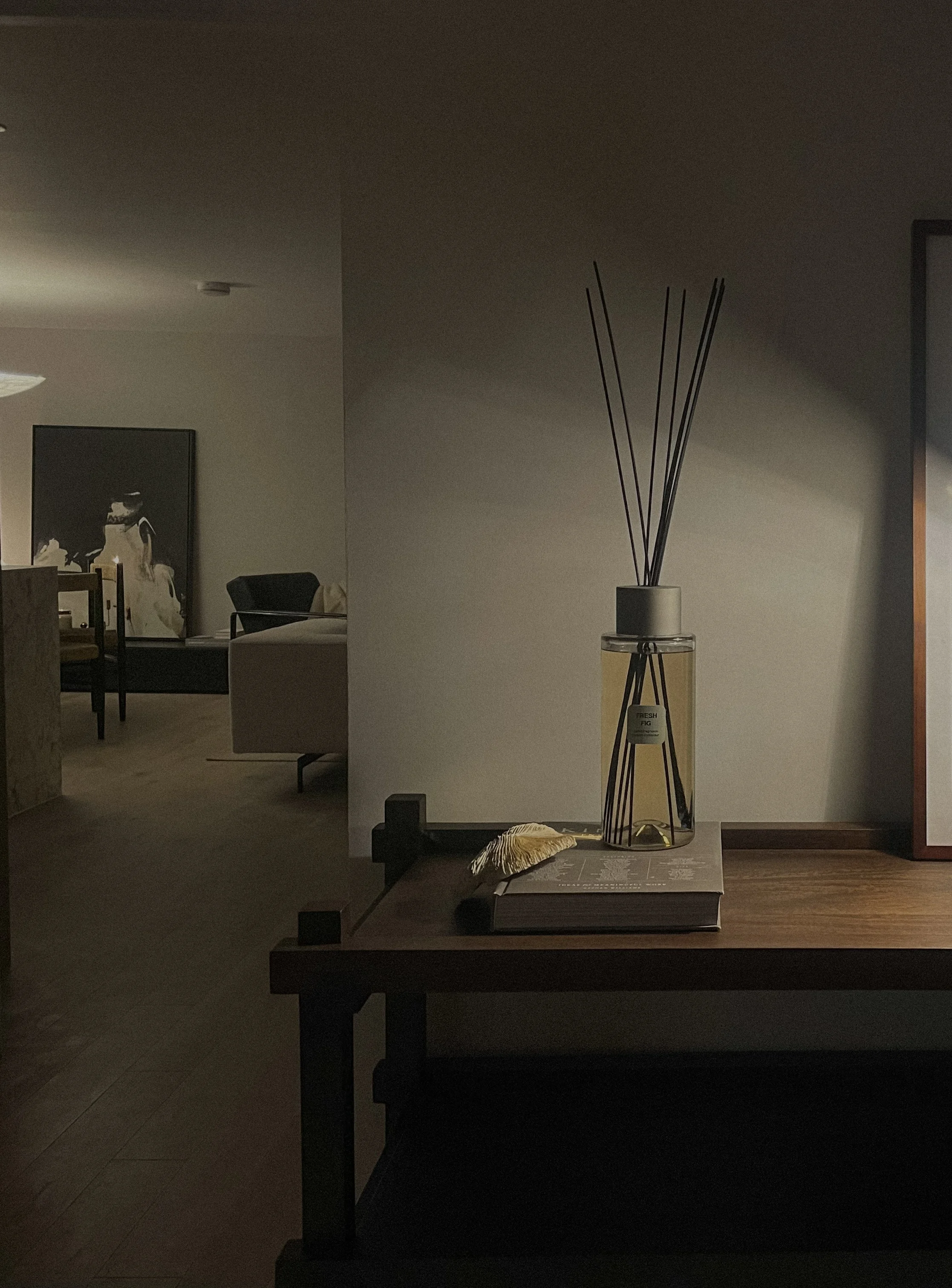 A close-up of a wooden table with a glass reed diffuser, a book, and a decorative feather. In the background, a living room with a sofa, chairs, and artwork on the wall is dimly visible.Residential interior design project in Scotland - Interior Desig