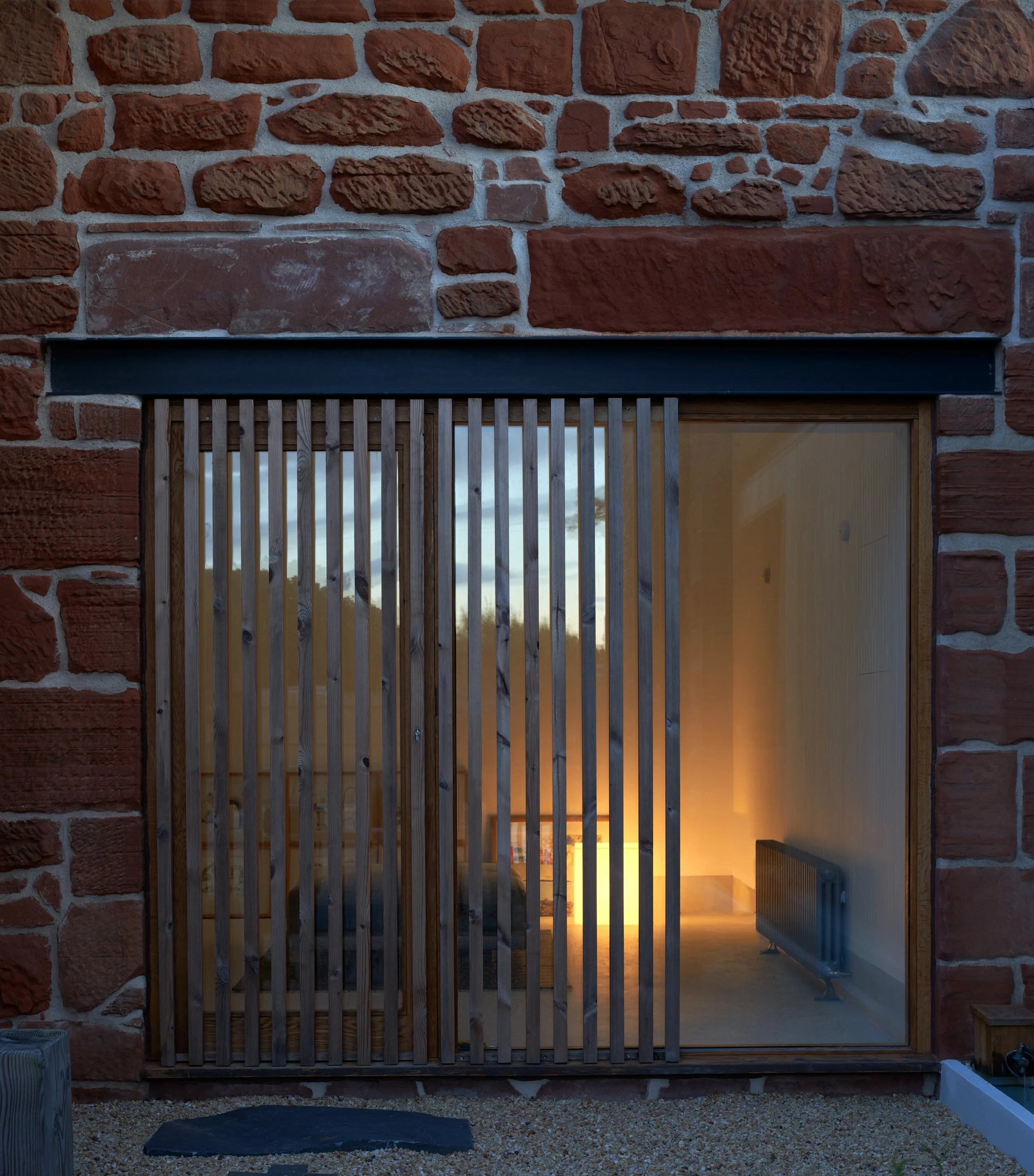 Exterior view of a building with a brick wall and a large glass window partly covered by wooden slats, showing a cozy interior with a yellow light, a radiator, and a view of the sky outside.Residential interior design project in Scotland - Interior D