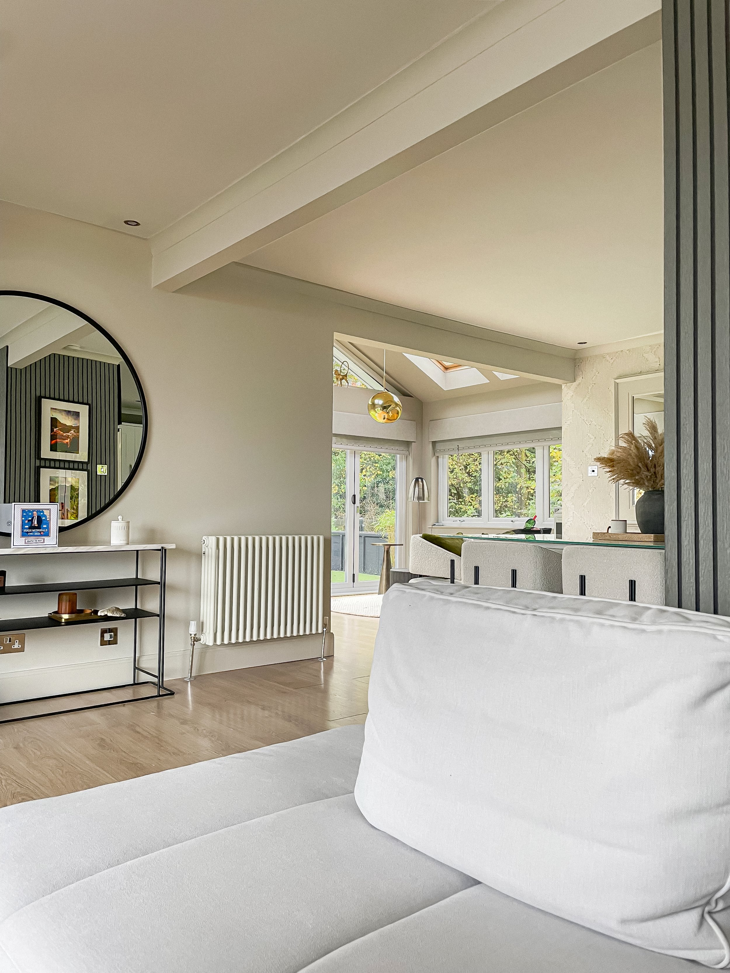 Modern living room with white sofa, wall-mounted mirror, black metal shelf, and view of patio doors leading outside, with plants visible through large windows.“Residential interior design project in Scotland by [Studio Name], working across Glasgow, 