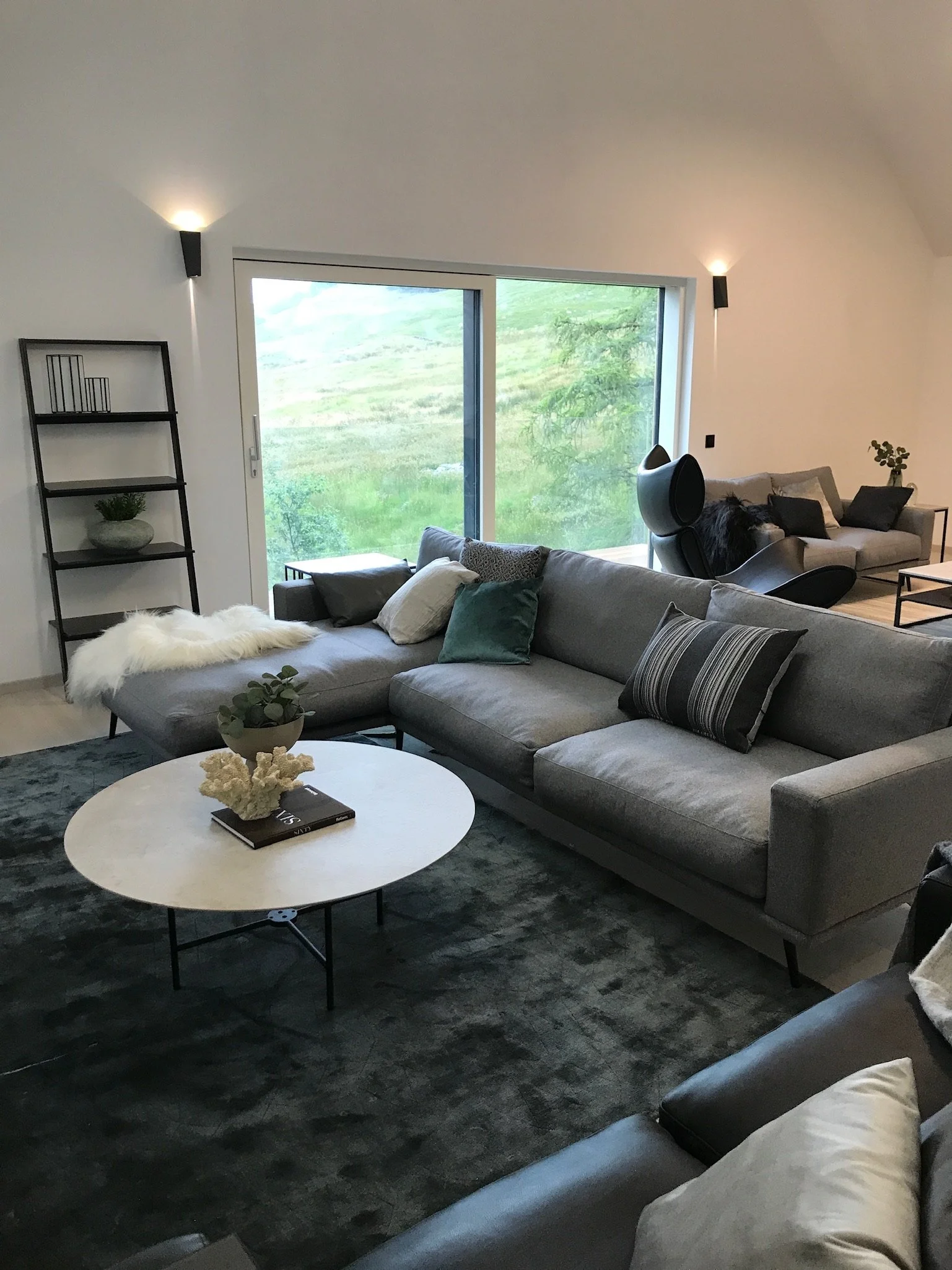 Modern living room with gray sectional sofa, black and gray pillows, a white round coffee table with a black book and decorative object, next to a black floor lamp, and a black shelving unit with decorative items. Large window showing green landscape