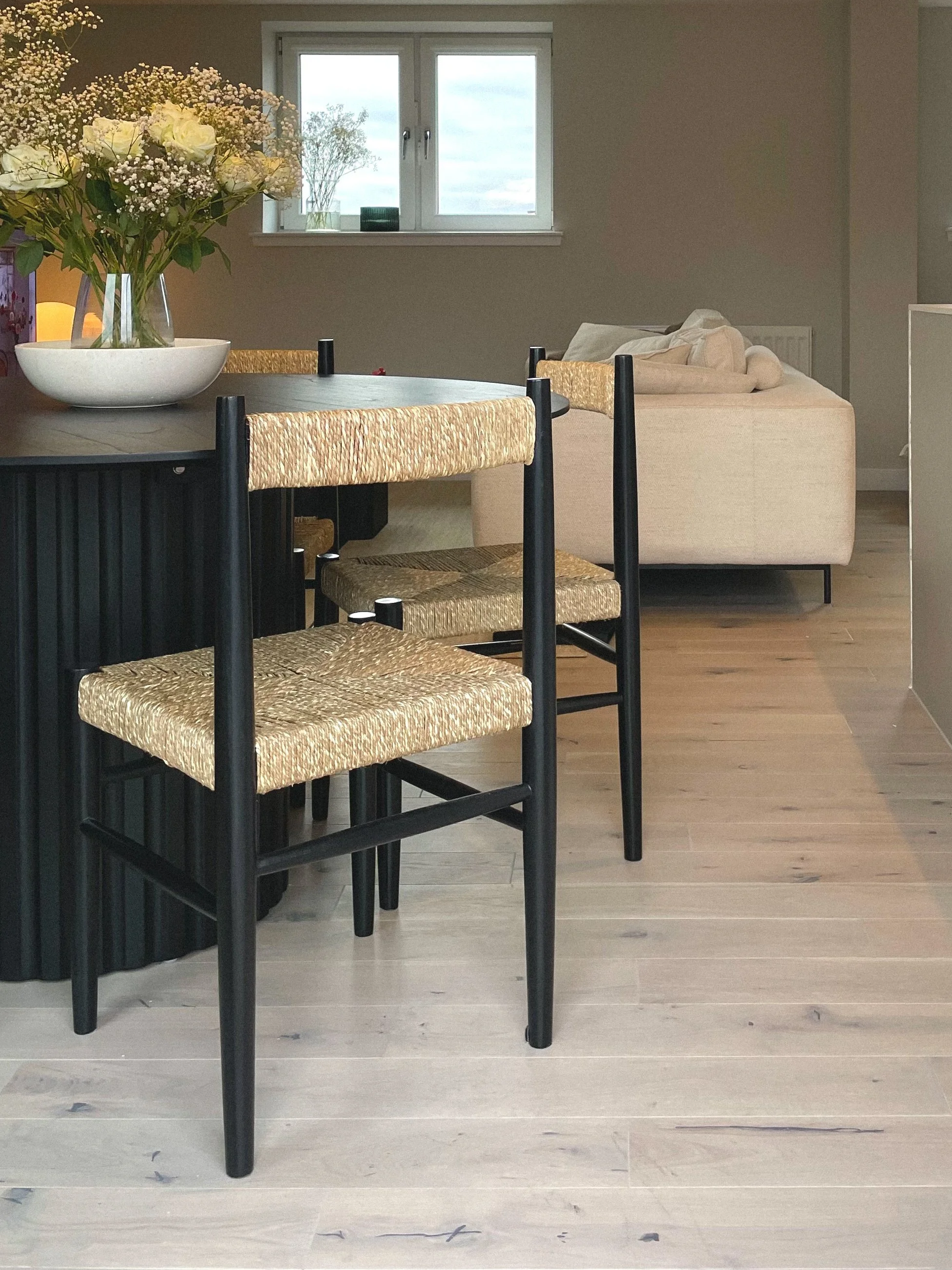 A dining area with a table and chairs, decorated with a large floral arrangement in a white bowl. In the background, a small sofa with light-colored cushions is visible, and a window shows an outside view of a cloudy sky.Residential interior design p