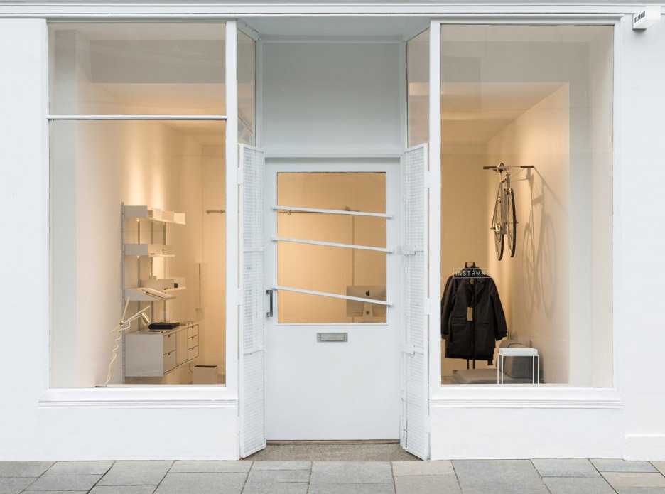 Minimalist storefront window display with white interior, a bicycle mounted on the right wall, a black jacket on a stand, and a small white stool, viewed through large glass windows and door.Residential interior design project in Scotland - Interior 