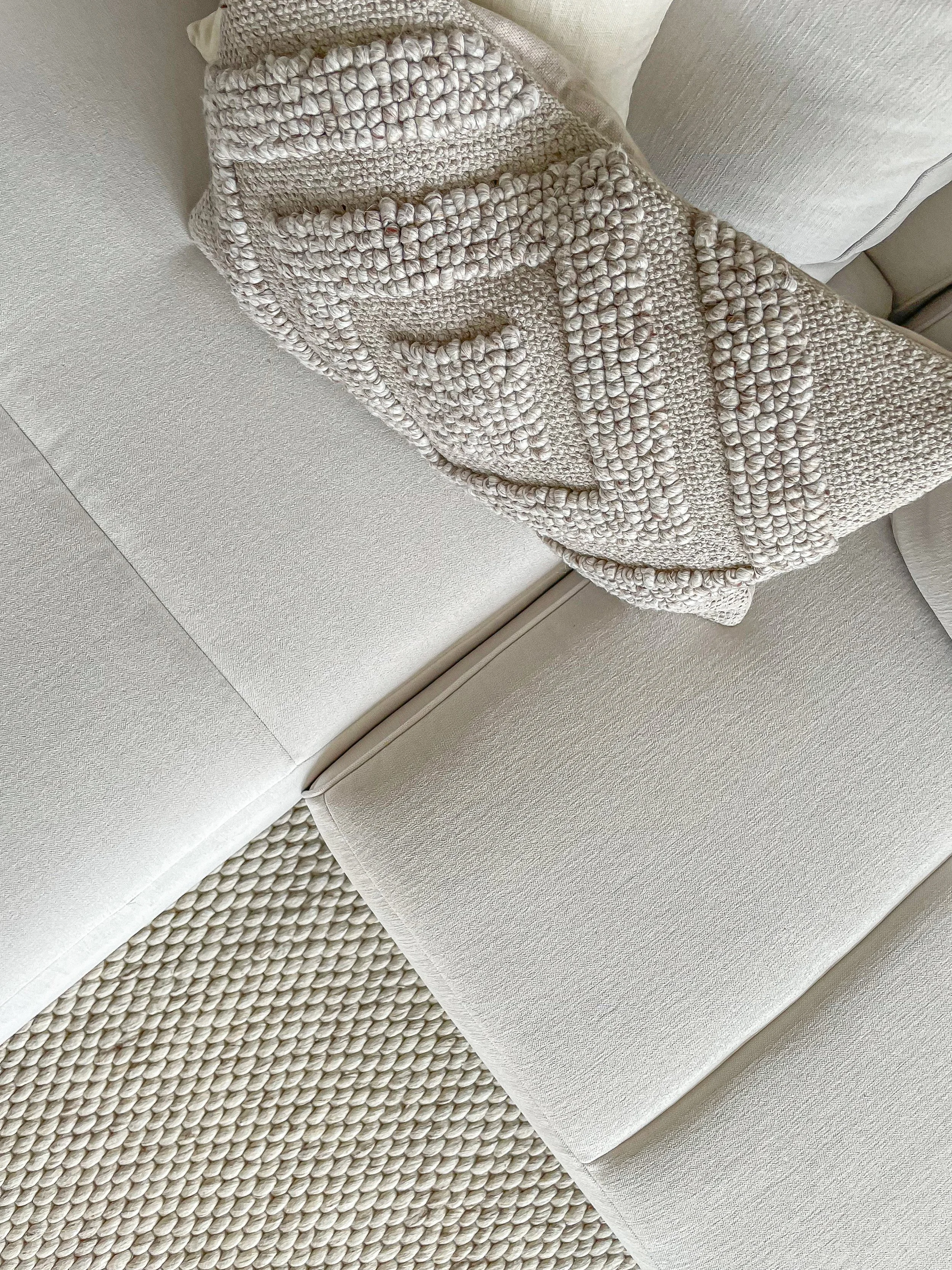 A beige textured couch with a cream knit blanket draped over the armrest, and a cream pillow on the side, placed on a matching textured rug.“Residential interior design project in Scotland by [Studio Name], working across Glasgow, Edinburgh, Dundee a