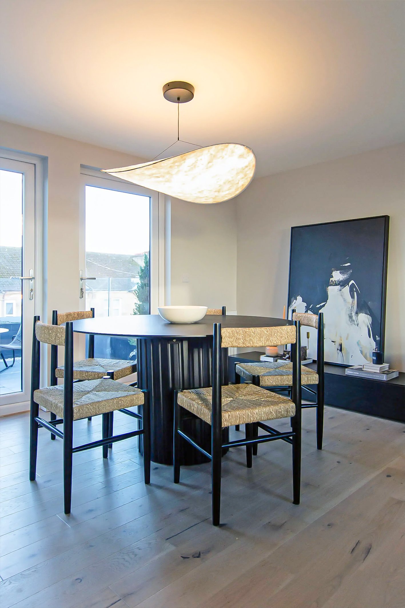 Modern dining room with a round black table, four woven chairs, a white bowl centerpiece, large windows, abstract wall art, and a ceiling light fixture.Residential interior design project in Scotland - Interior Designer Scotland - Interiors 