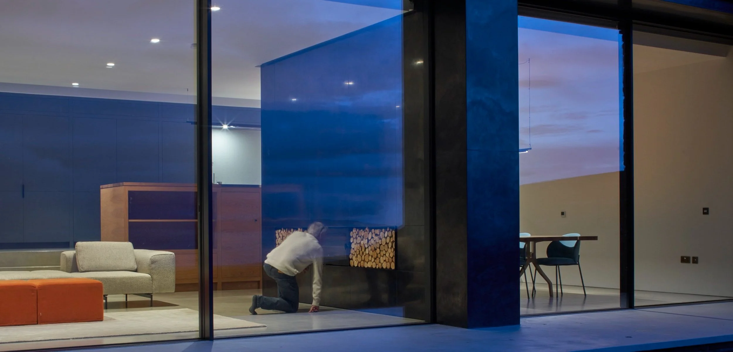 Person kneeling inside a modern home with large glass windows, a sitting area with a gray couch and orange ottoman, and a dining area with a table and chairs, during dusk or evening.“Residential interior design project in Scotland by [Studio Name], w