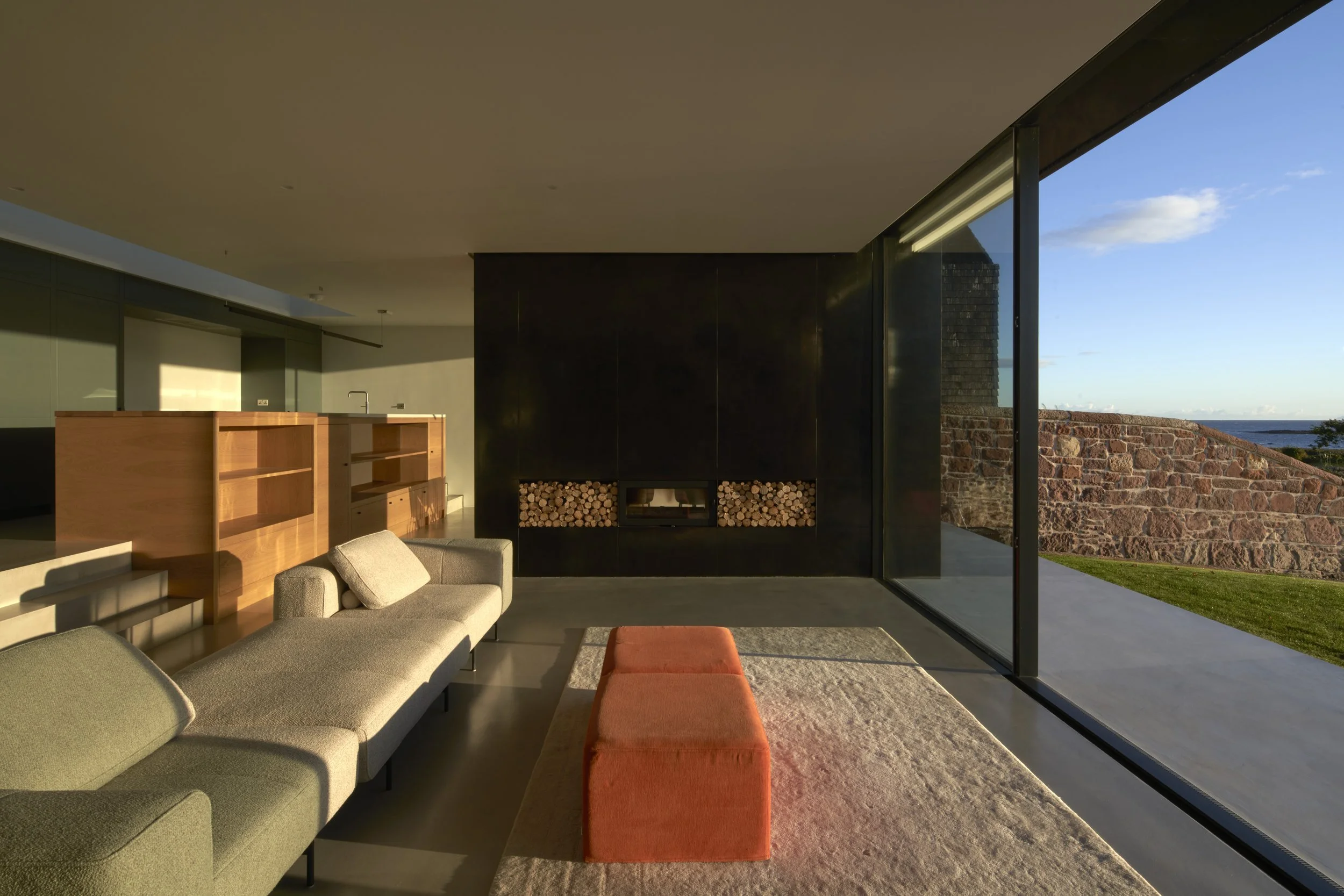 Modern living room with beige and green sofas, orange ottoman, wood and black fireplace, large glass window showing stone wall and outside greenery, and sunlight streaming in.“Residential interior design project in Scotland by [Studio Name], working 