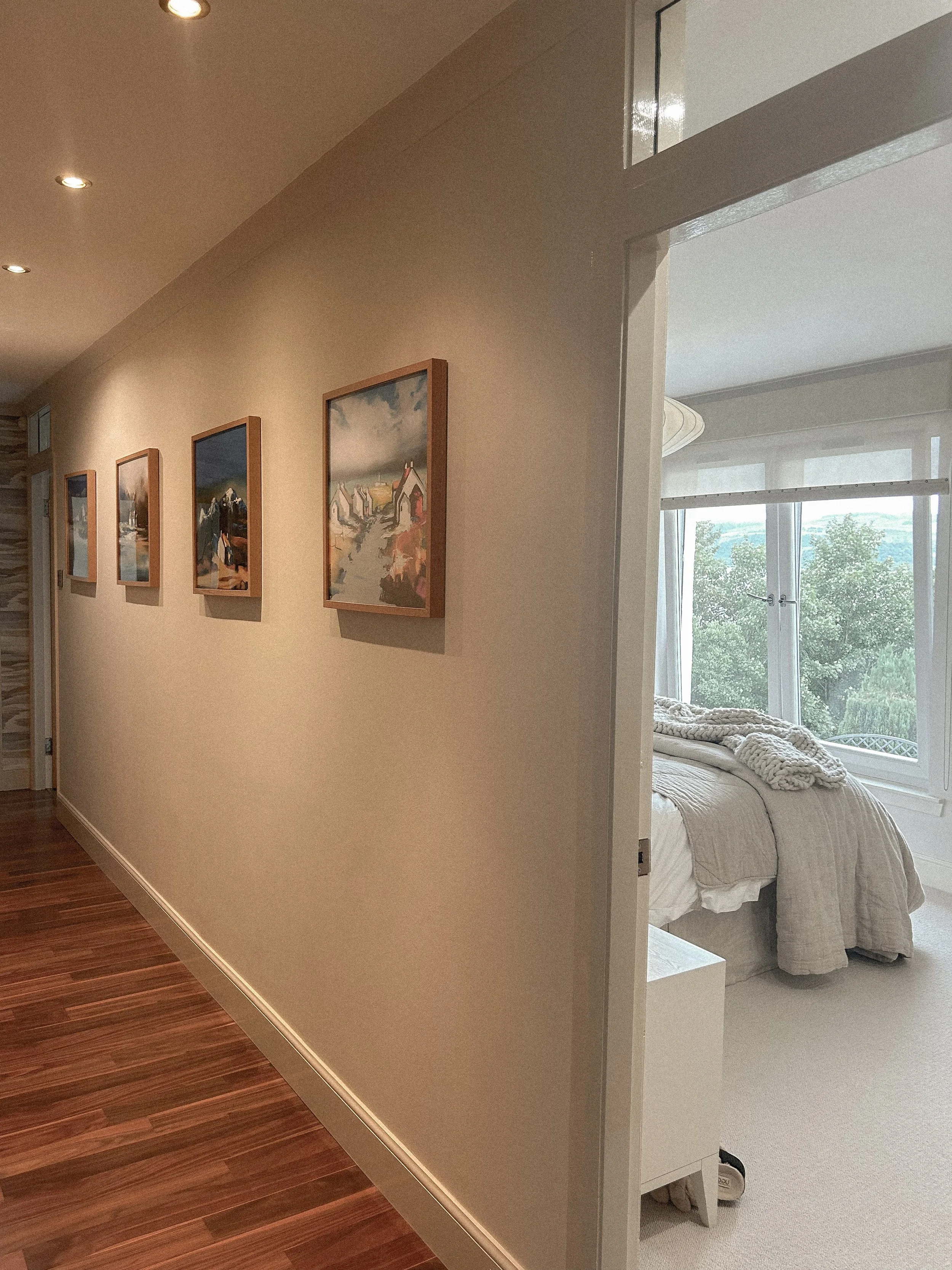 A hallway with wooden flooring and five framed landscape paintings on the wall. To the right, an open door reveals a bedroom with large windows, a bed with light-colored linens, and a view of trees outside.“Residential interior design project in Scot
