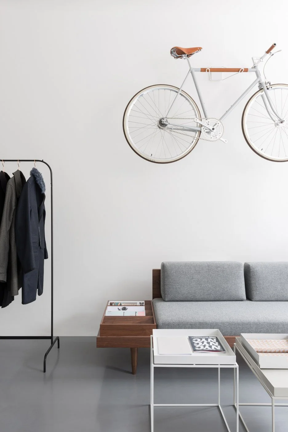 Minimalist interior with a white wall, a mounted white bicycle with brown accents, a gray sofa, a small white table with magazines, a wooden side table, and a clothing rack with jackets.Residential interior design project in Scotland - Interior Desig