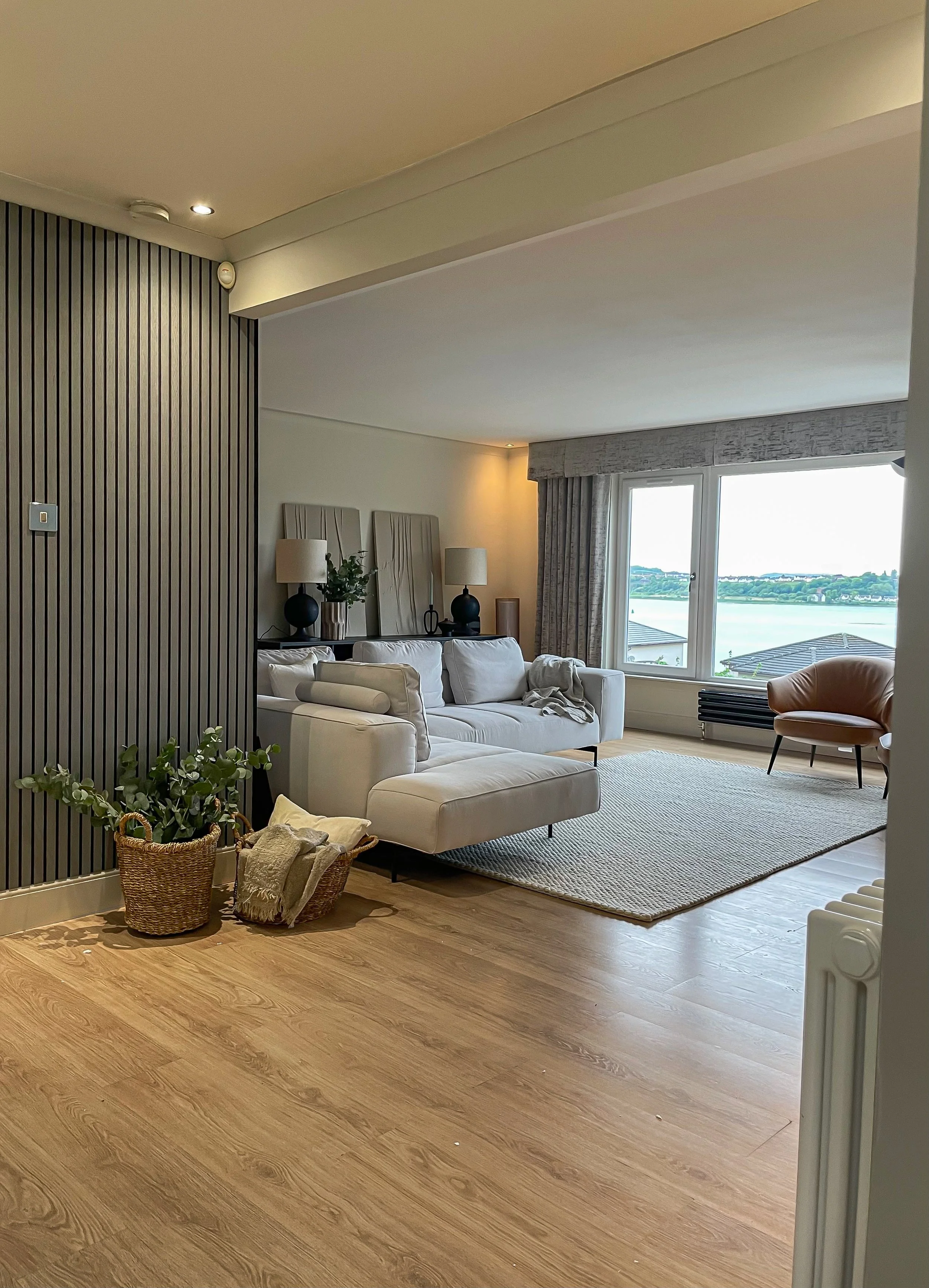 Modern living room with white sofa, armchair, large window overlooking water, decorative lamps, and potted plant.“Residential interior design project in Scotland by [Studio Name], working across Glasgow, Edinburgh, Dundee and the Highlands.”

“Contem