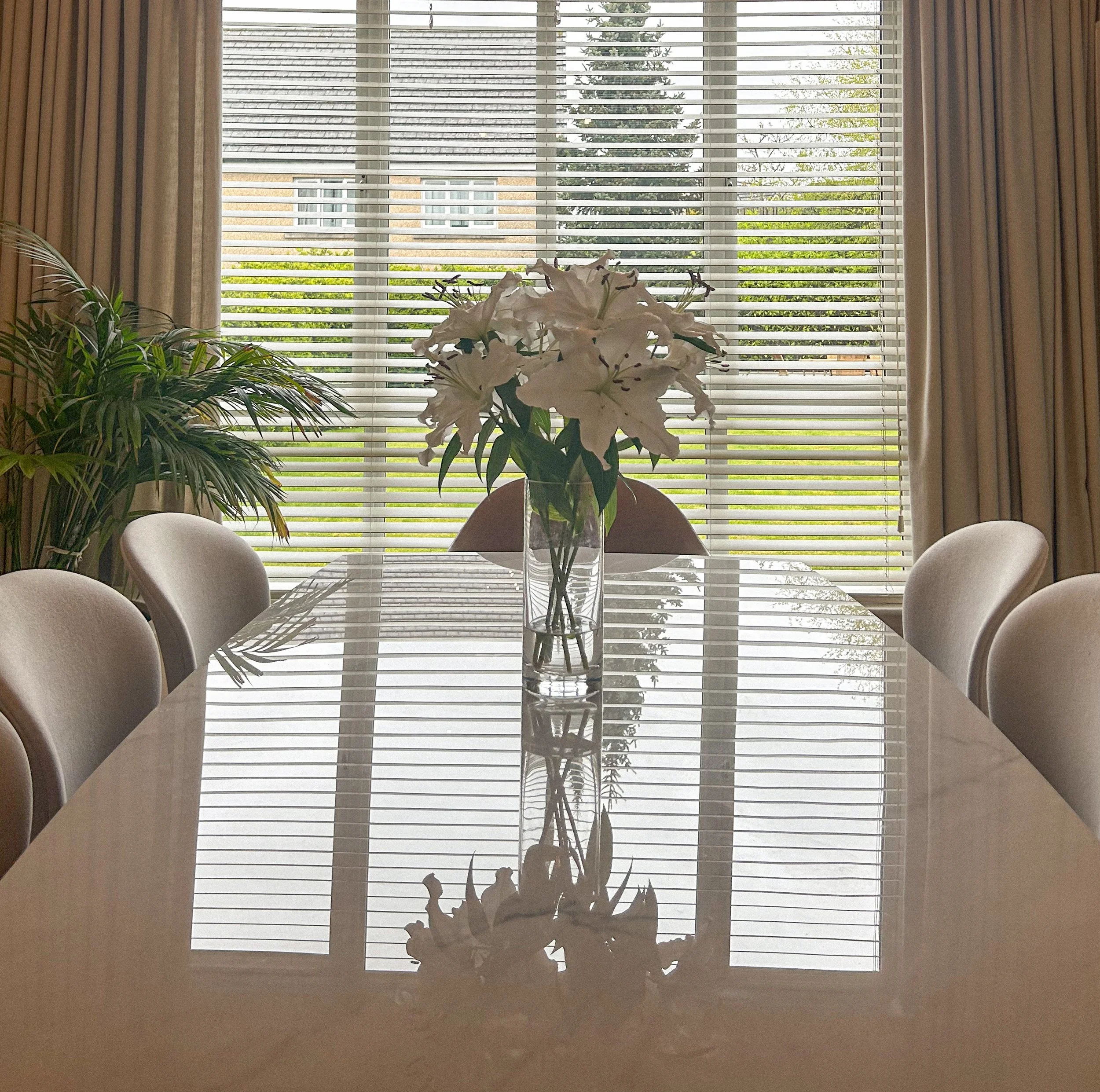 A dining room with a glass table, six chairs, a vase of white lilies, a potted plant, and window blinds showing a garden outside.Residential interior design project in Scotland - Interior Designer Scotland - Interiors 
