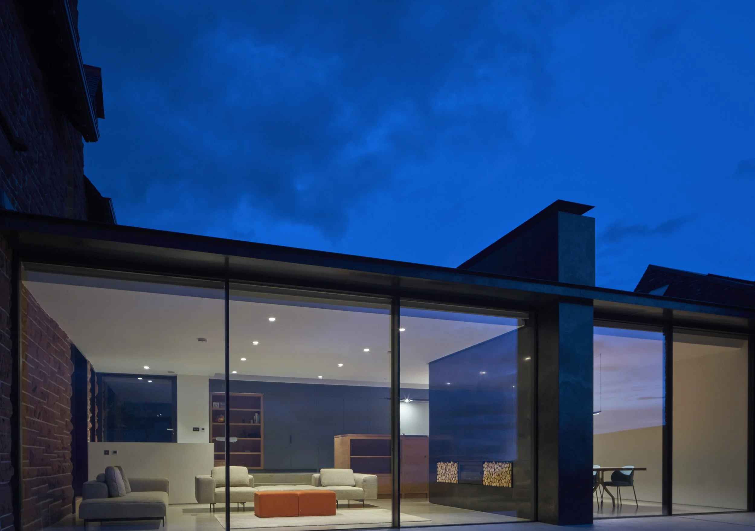 Modern house with large glass windows, illuminated interior showing living room with beige sofas and orange ottoman, and a dining area with a table and chairs, against a twilight sky.“Residential interior design project in Scotland by [Studio Name], 