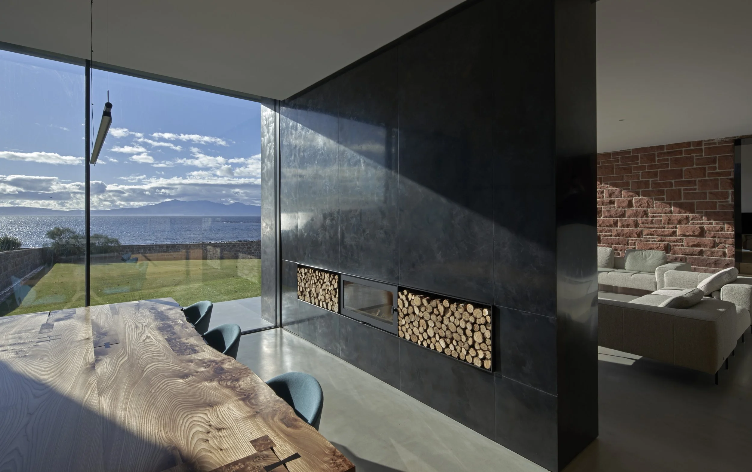 Modern living room with large glass window overlooking a lake and mountains, featuring a wooden dining table, black grid wall with stacked firewood, and white sectional sofa.Residential interior design project in Scotland - Interior Designer Scotland