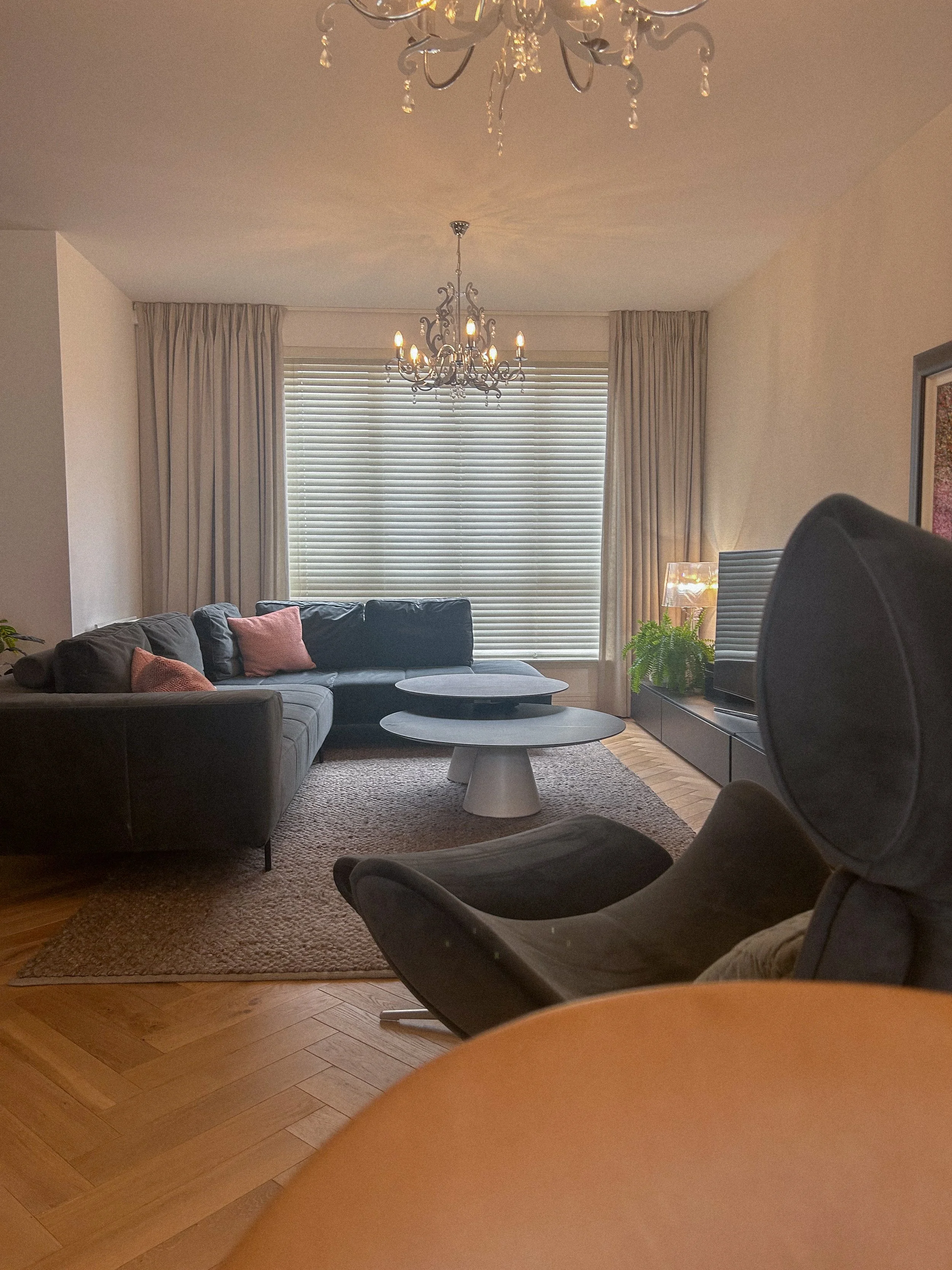 Living room with black sectional sofa with pink pillows, two round coffee tables, a TV on a low TV stand, a potted plant, a tall lamp, a window with closed blinds and curtains, two chandeliers hanging from the ceiling, and hardwood flooring with a ru