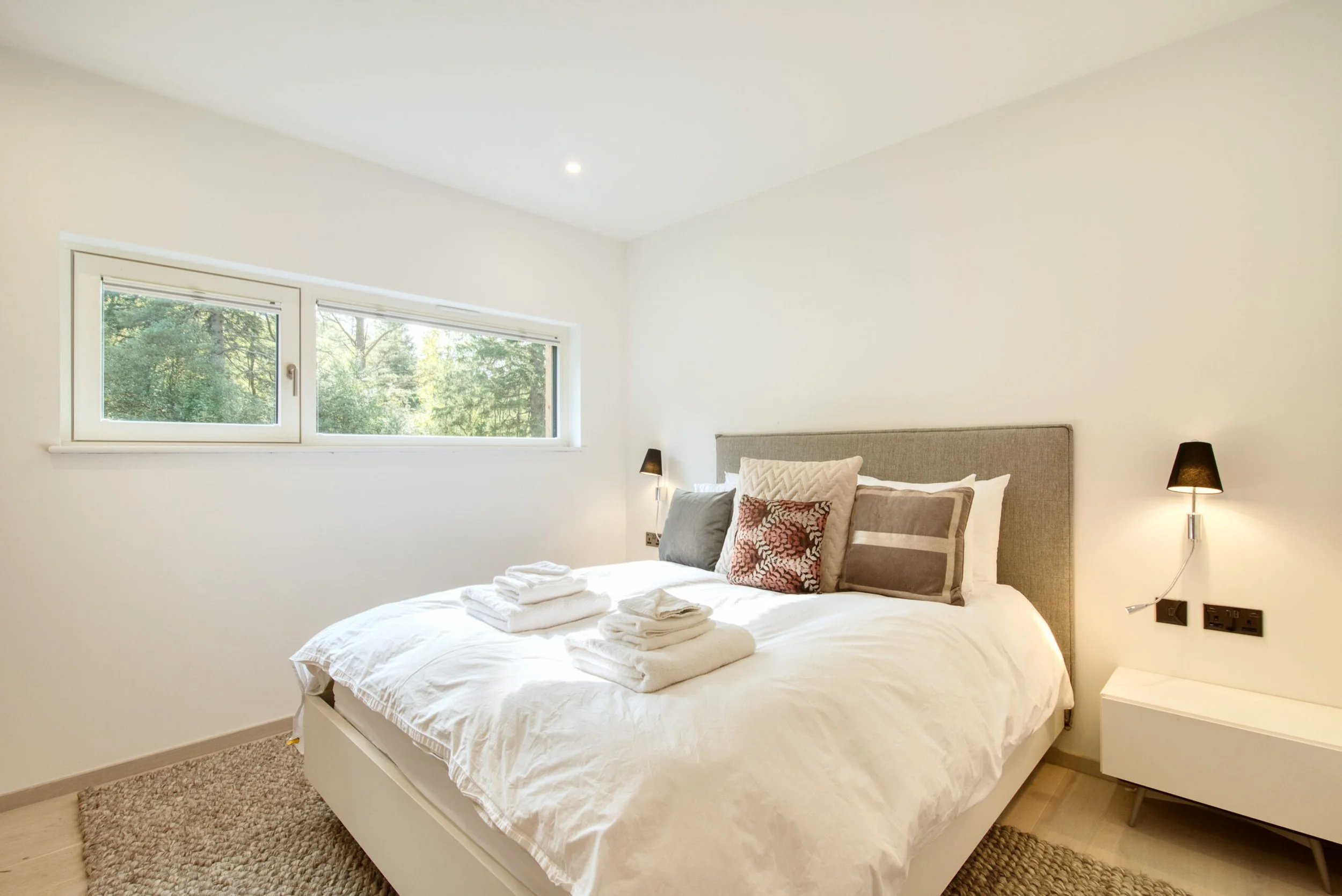 Bedroom with a large bed, white bedding, multiple pillows, window overlooking green trees, wall-mounted lamps, and a white nightstand.Residential interior design project in Scotland - Interior Designer Scotland - Interiors 