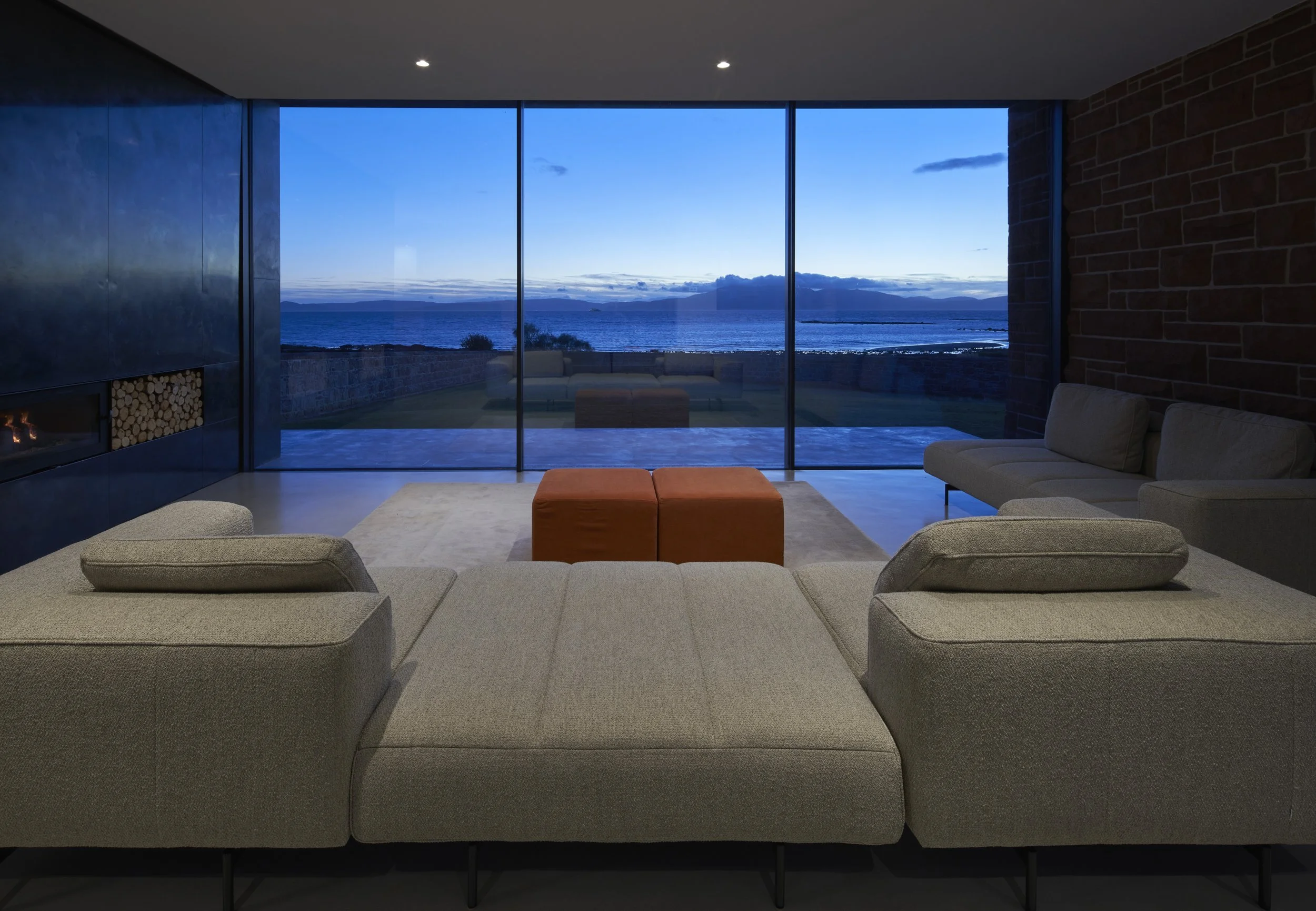 Modern living room with large floor-to-ceiling windows overlooking a body of water at dusk. The room has beige sectional sofas, an orange ottoman, and a fireplace on the left wall.“Residential interior design project in Scotland by [Studio Name], wor