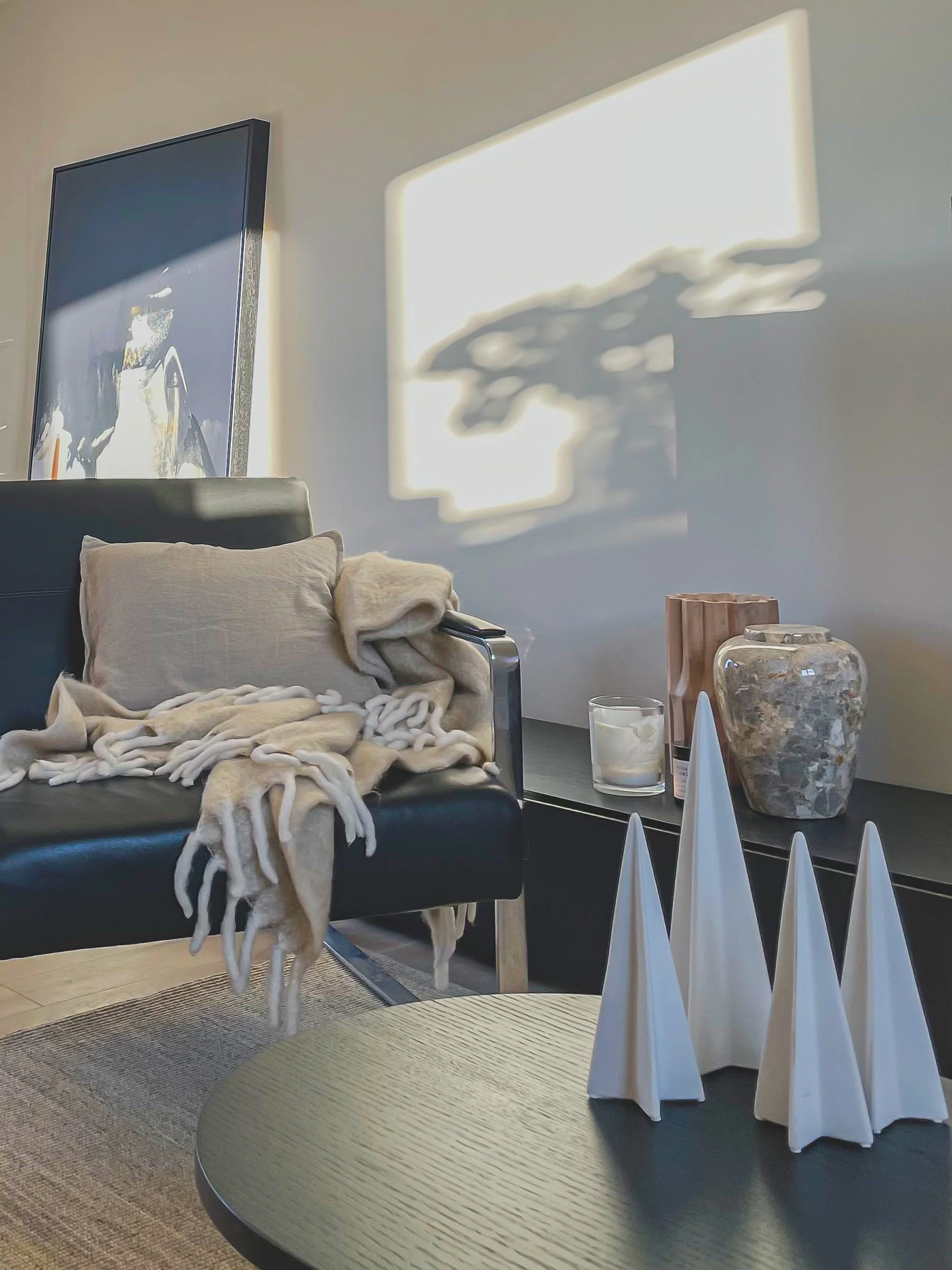 Living room with a black sofa, beige pillows, and a beige throw blanket, a black side table holding a candle, a decorative vase, and a pink sculpture, a round beige coffee table with white decorative objects, and sunlight casting shadows on the wall.