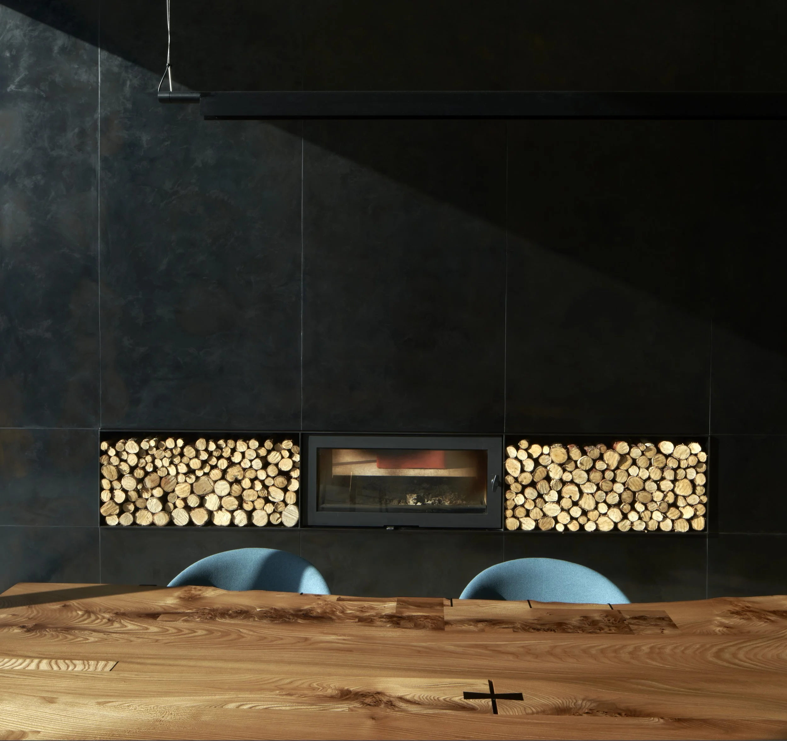 Interior of a modern room showing a black tiled wall with a built-in fireplace flanked by stacked firewood, a wood table in the foreground, and two teal chairs.“Residential interior design project in Scotland by [Studio Name], working across Glasgow,