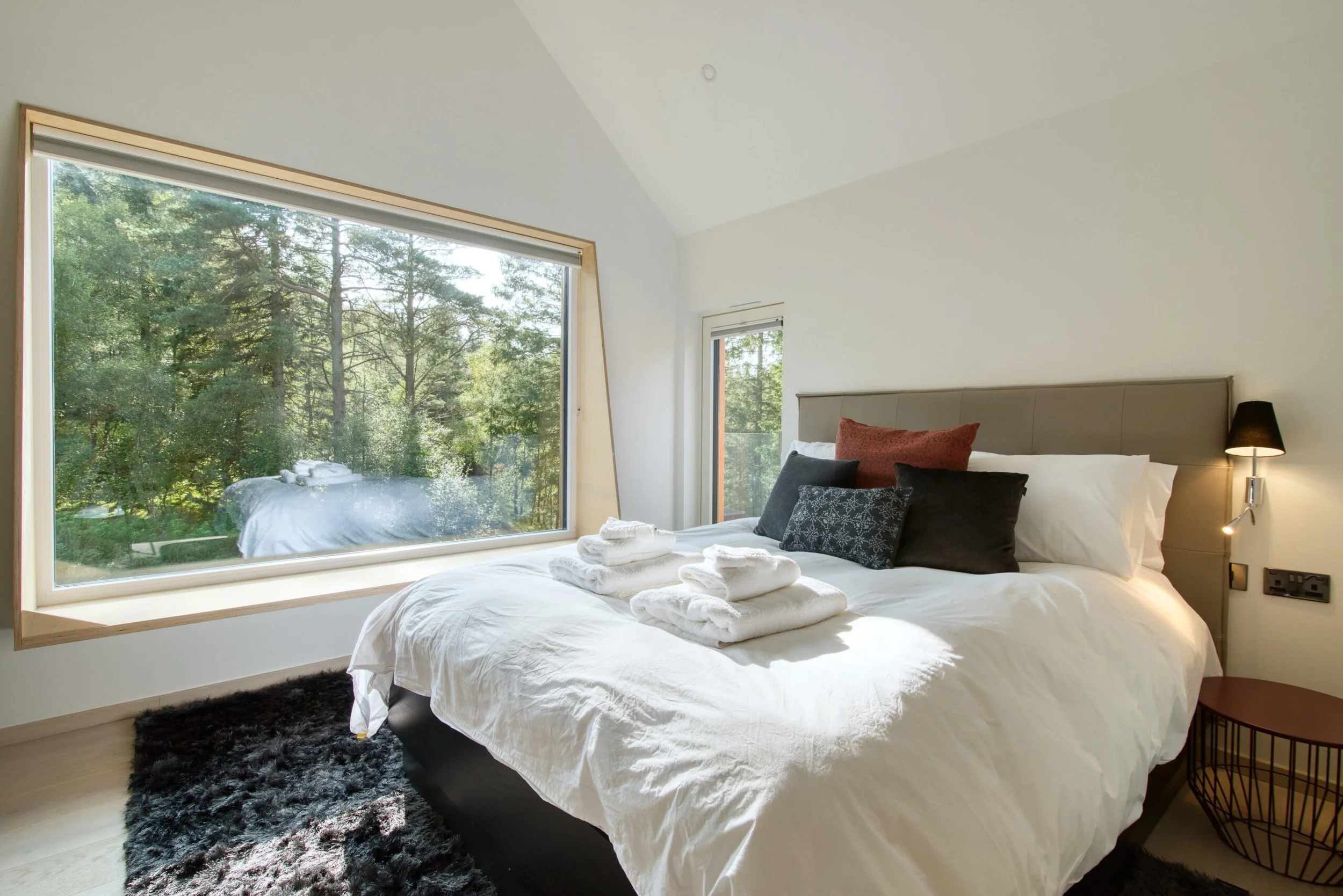 A bedroom with a large window showing a forest view, a bed with white linens, and pillows, with towels on top.Residential interior design project in Scotland - Interior Designer Scotland - Interiors 
