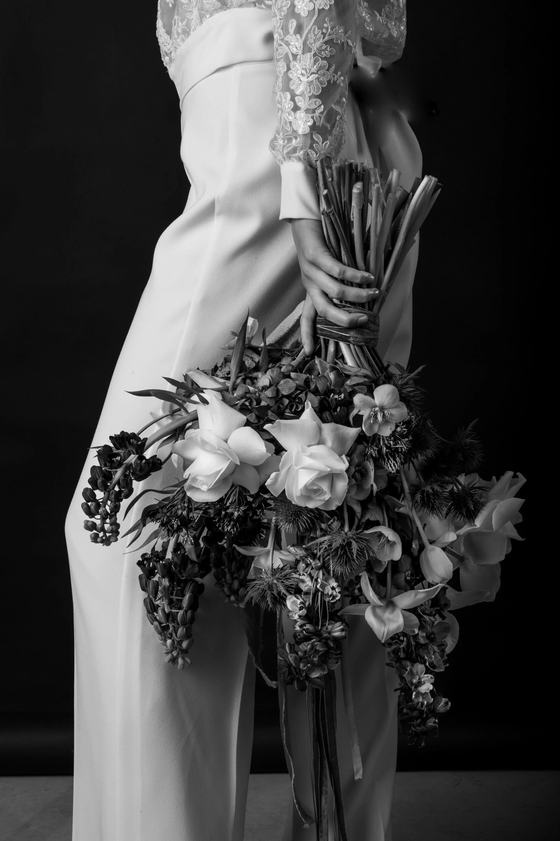 Black and white photo of a person holding a bouquet of flowers against a dark background. The person is dressed in a lace top and white pants.
