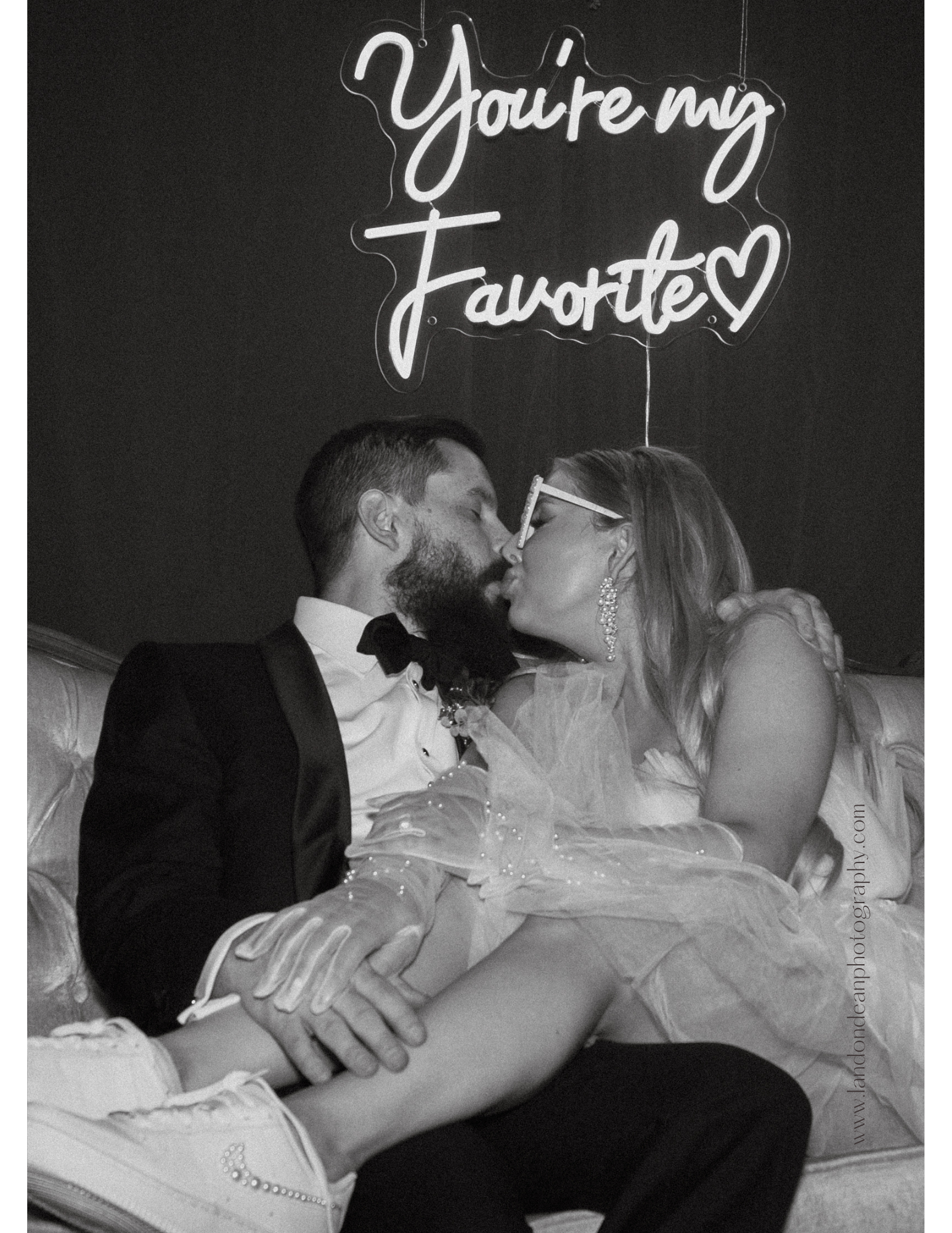 Black and white photo of a couple kissing on a couch, with a neon sign reading "You're my Favorite." The woman is wearing a tulle dress, and both are dressed semi-formally with relaxed footwear. The image has a romantic and candid feel.