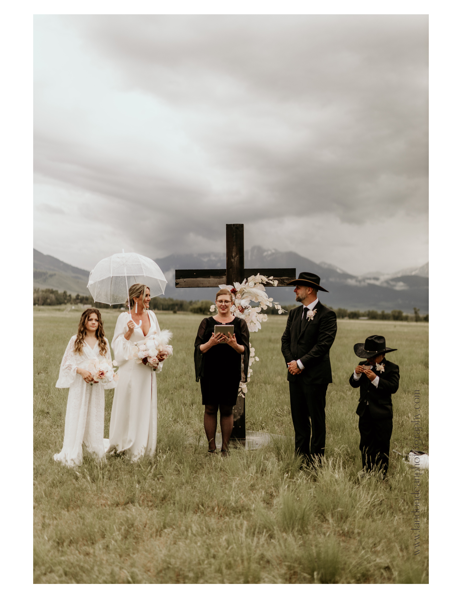 Outdoor wedding ceremony with bride in white holding an umbrella, a groom in a suit and cowboy hat, a young girl in a white dress, an officiant, and a young boy in a suit and cowboy hat, near a wooden cross in a grassy field with cloudy skies.
