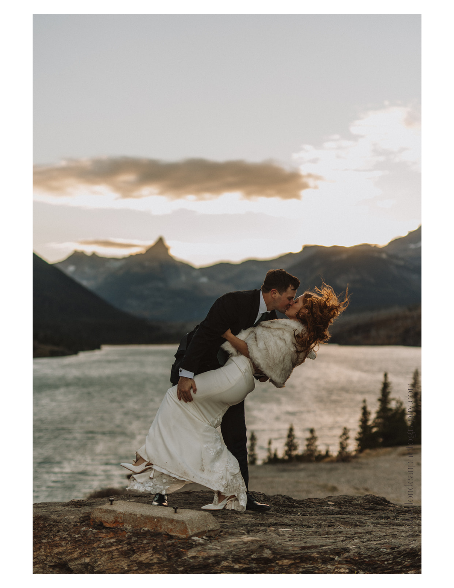 Bride and groom kiss on a rocky cliff with a lake and mountains in the background at sunset.