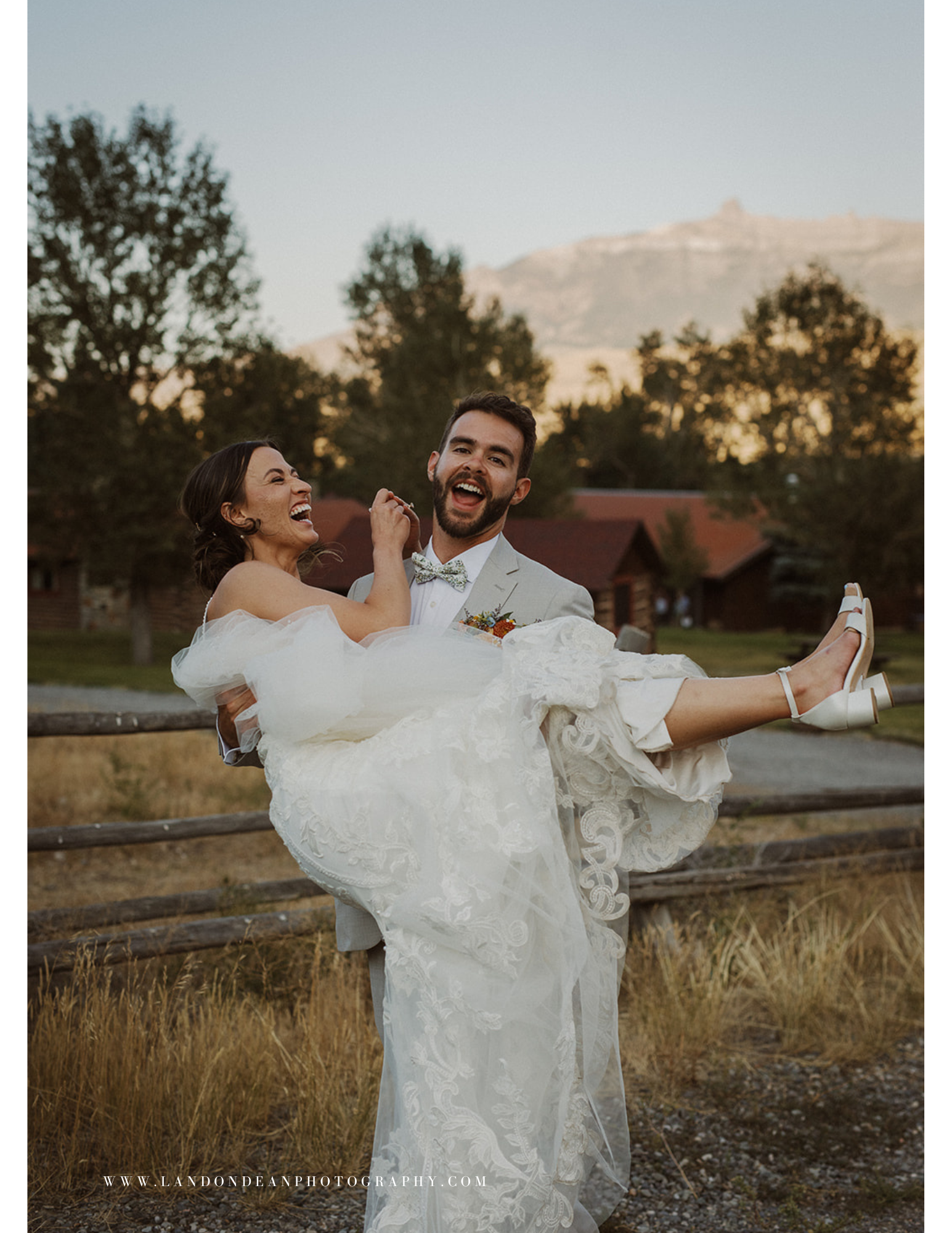 Bride and groom laughing in outdoor wedding setting, with groom carrying bride by a wooden fence and trees, mountains in background.