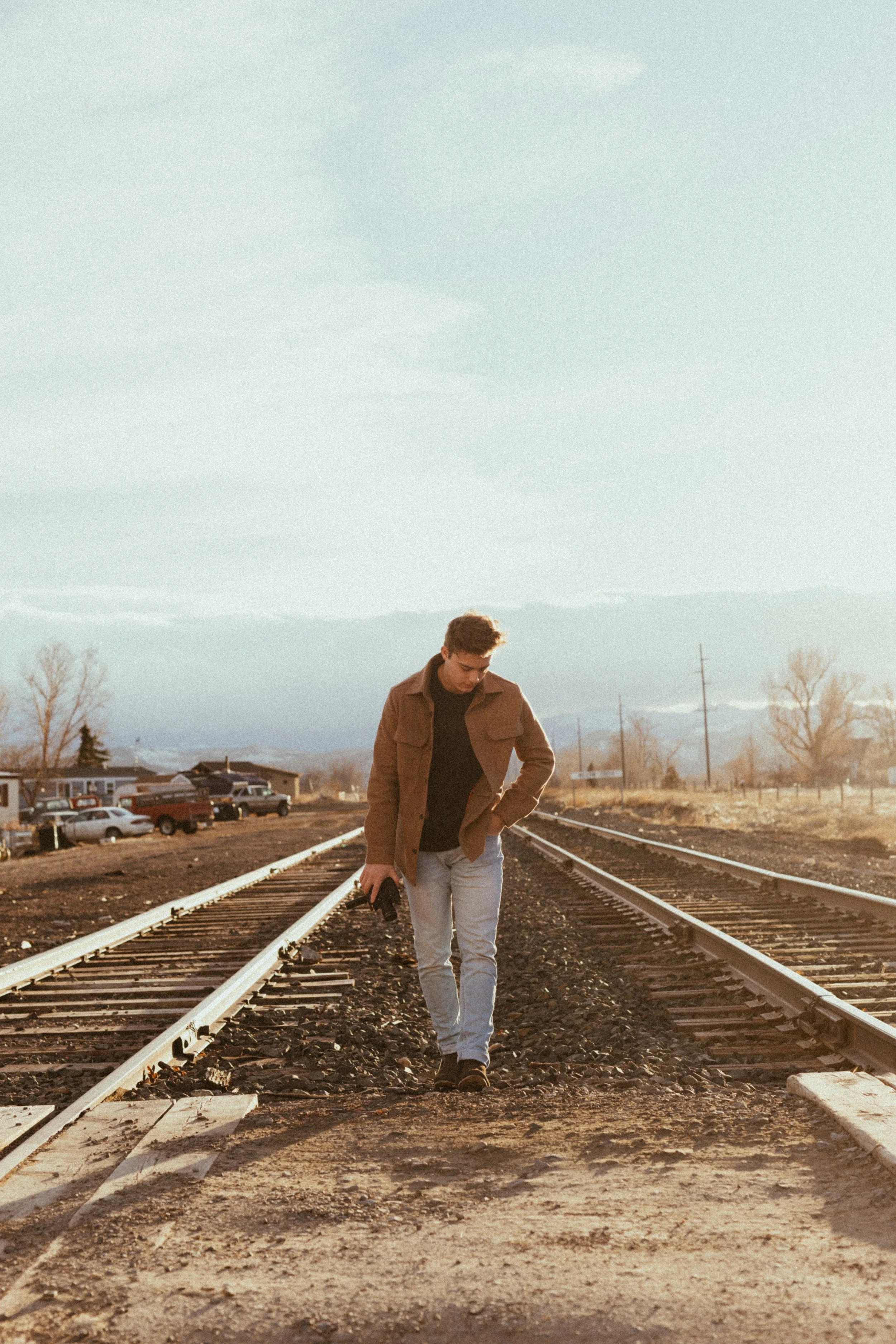 Landon Dean Photography walking on railroad tracks in a rural area, holding a camera, wearing a brown jacket and jeans, with cars and houses in the background.