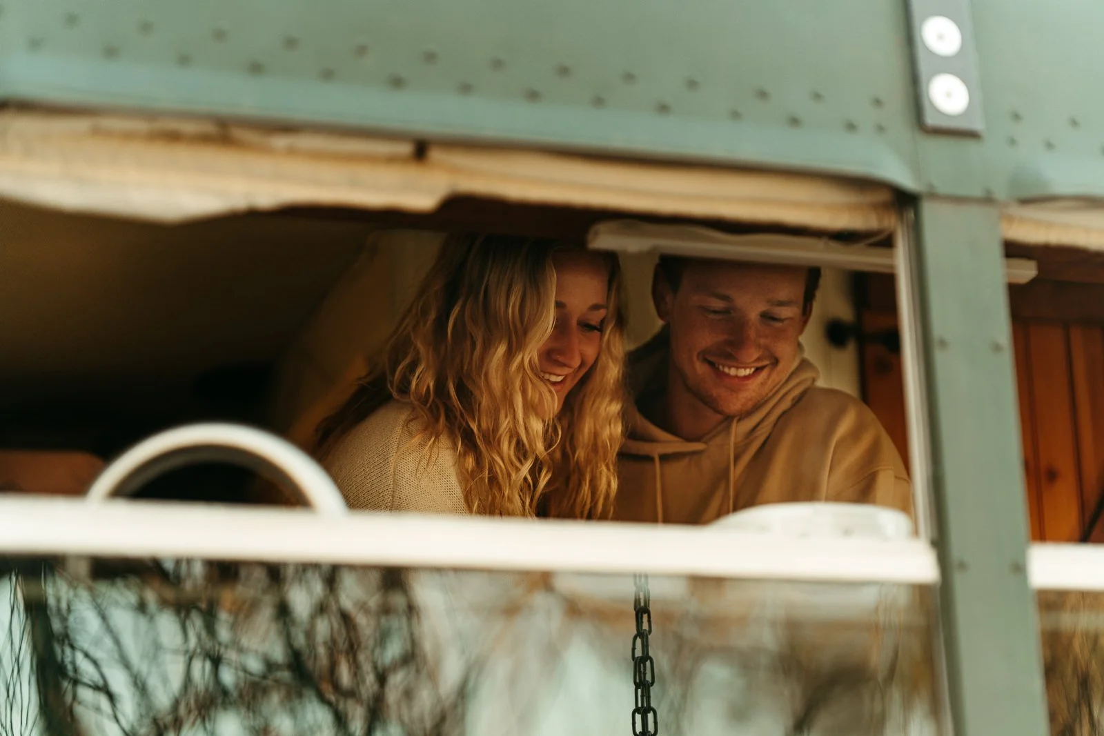 Nikki and Sawyer smiling as they make breakfast together in their skoolie, visible from a bus window.