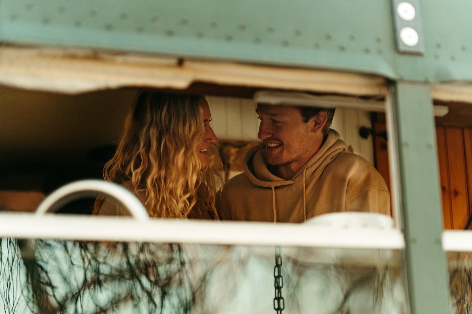 Nikki and Sawyer smile at each other while they make breakfast together in their skoolie, visible from a bus window.
