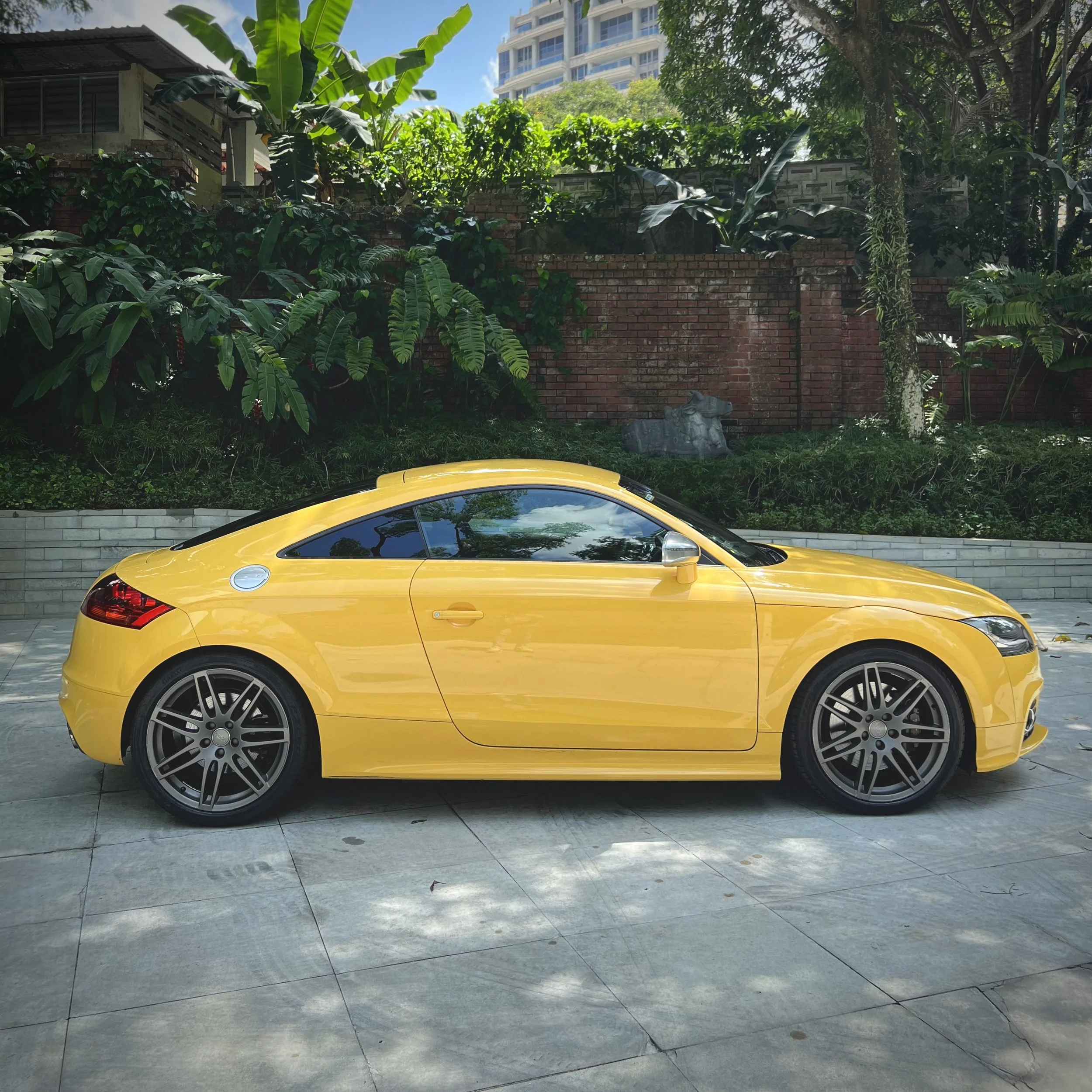 Yellow sports car parked on a stone pavement with lush green foliage in the background.