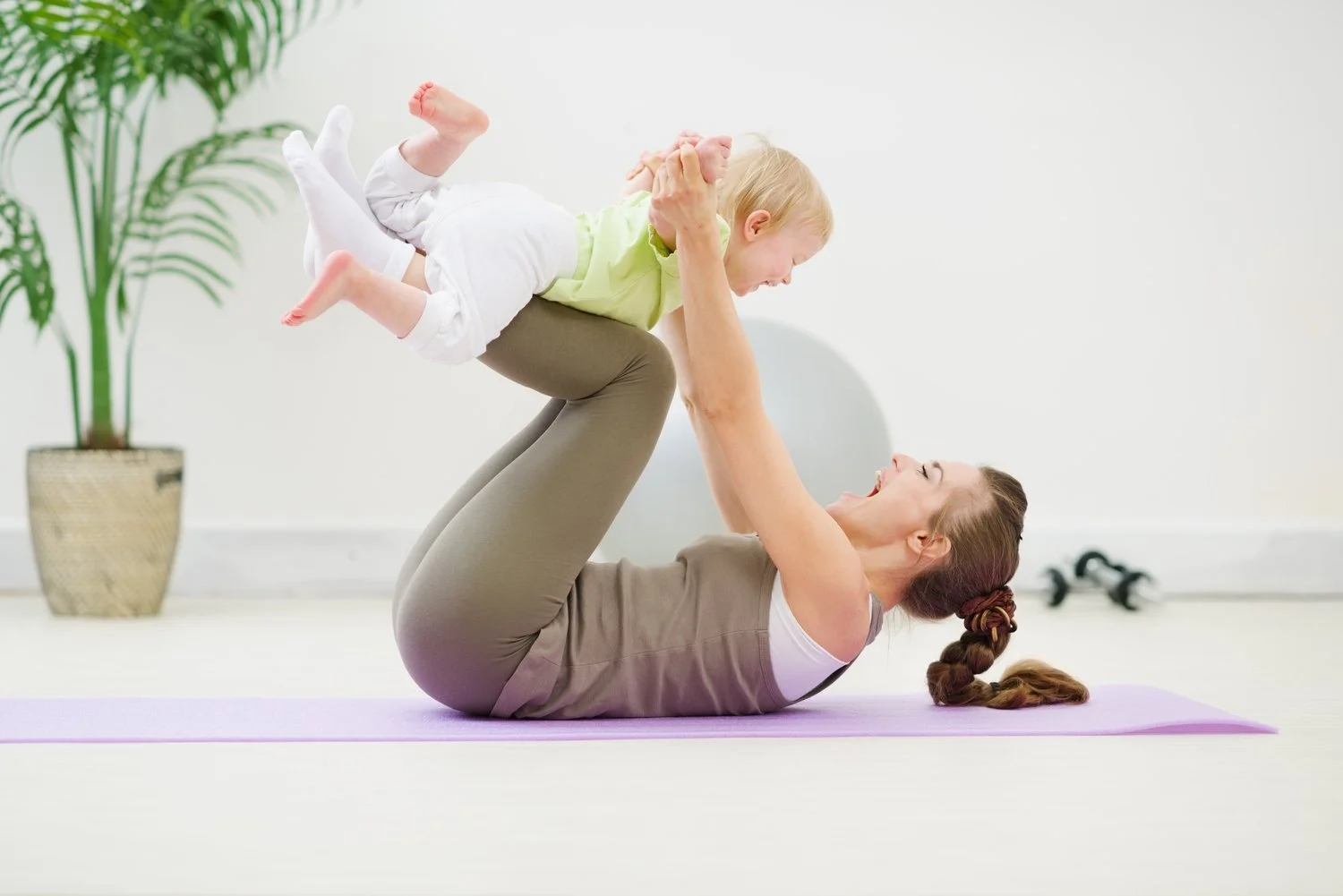Joyful mother in a yoga pose lifting her baby in the air, enjoying a playful moment together on a yoga mat in a serene indoor environment
