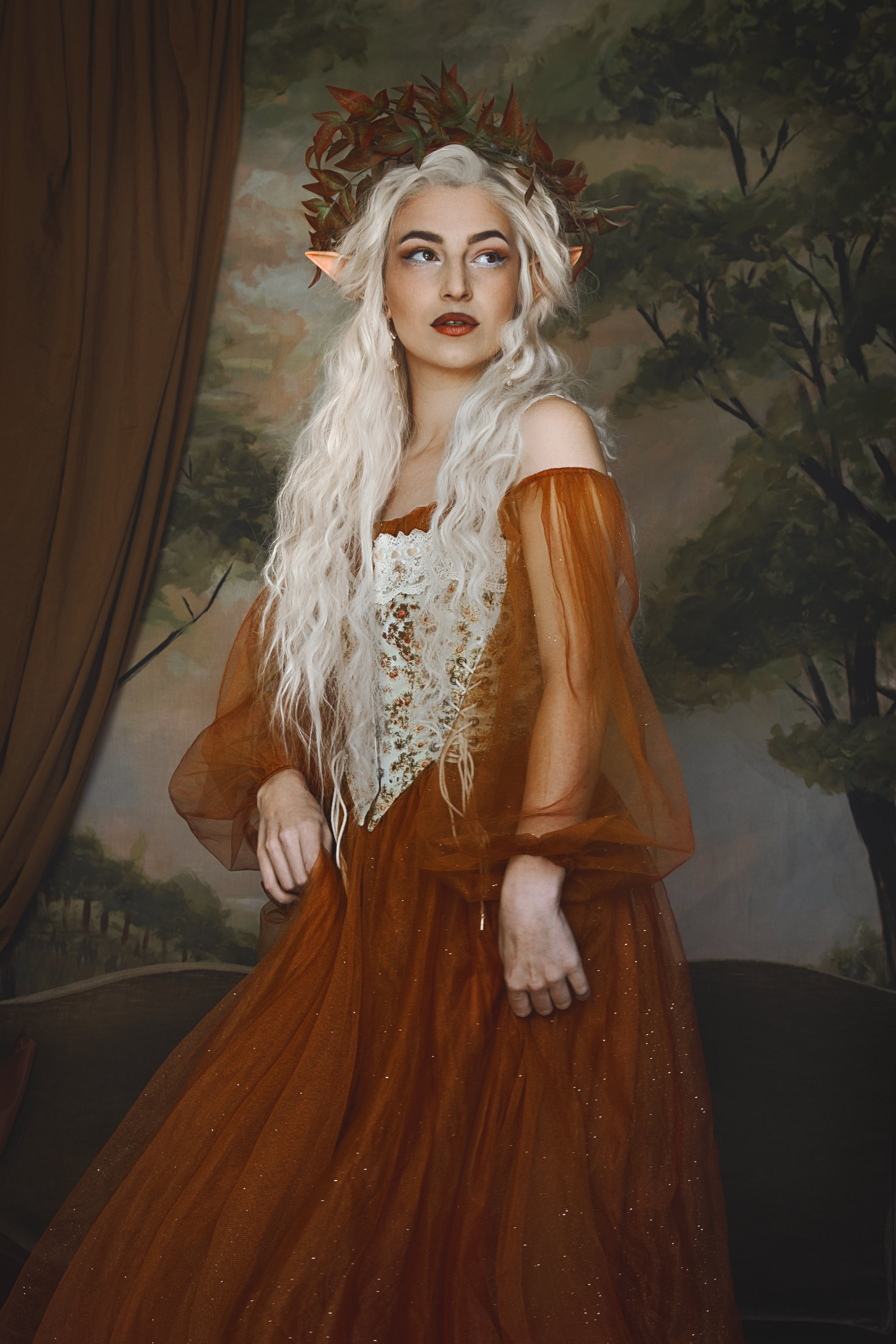 Woman dressed as a fantasy character with long white hair, wearing a brown and white gown, with a leafy headpiece, standing against a backdrop of trees.