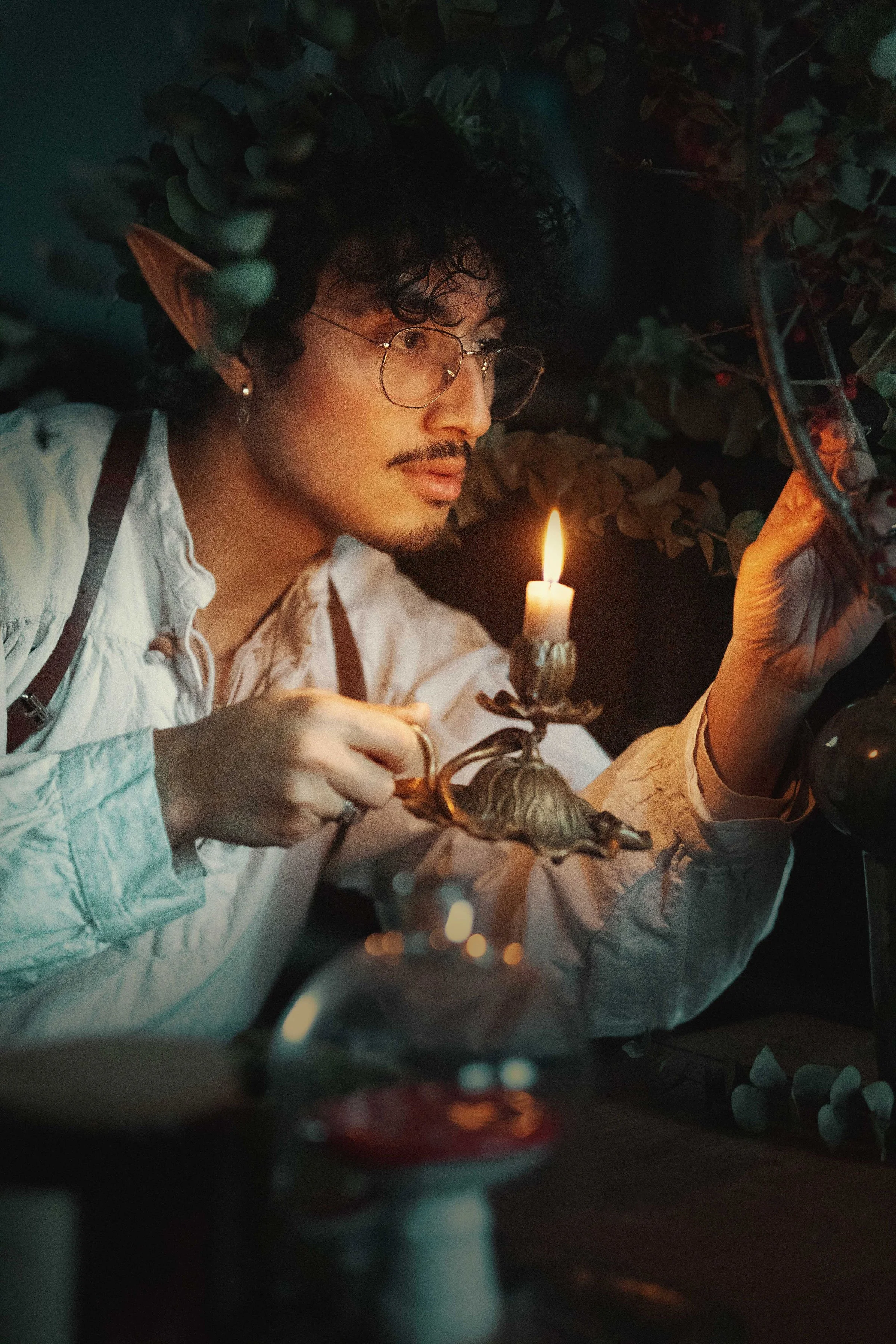Dreamy fine art portrait of Dewa as a folkloric alchemist, surrounded by glass bottles, herbs and candlelight. Painterly fantasy photography inspired by historical apothecaries and storytelling portraiture. Studio shoot in the Netherlands.