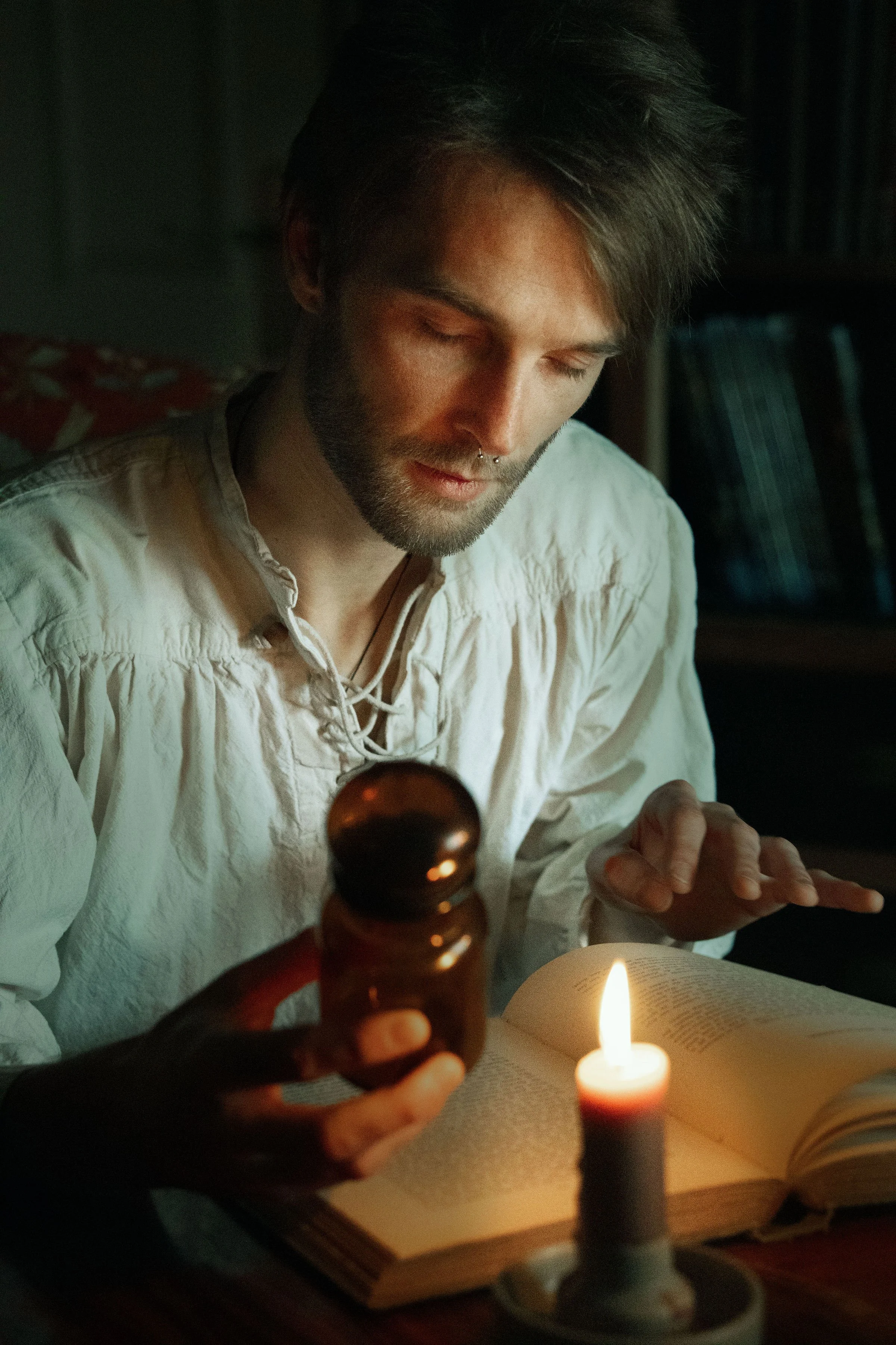 Dreamy fine art portrait of Rico as a scholar reading by candlelight. Painterly fantasy photography inspired by folklore and classical paintings, featuring books, glass bottles and warm light. Moody, romantic storytelling imagery in a fine art studio