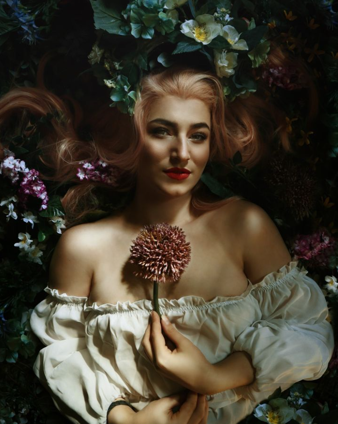 Woman lying on bed of flowers, holding a chrysanthemum, wearing an off-shoulder blouse.