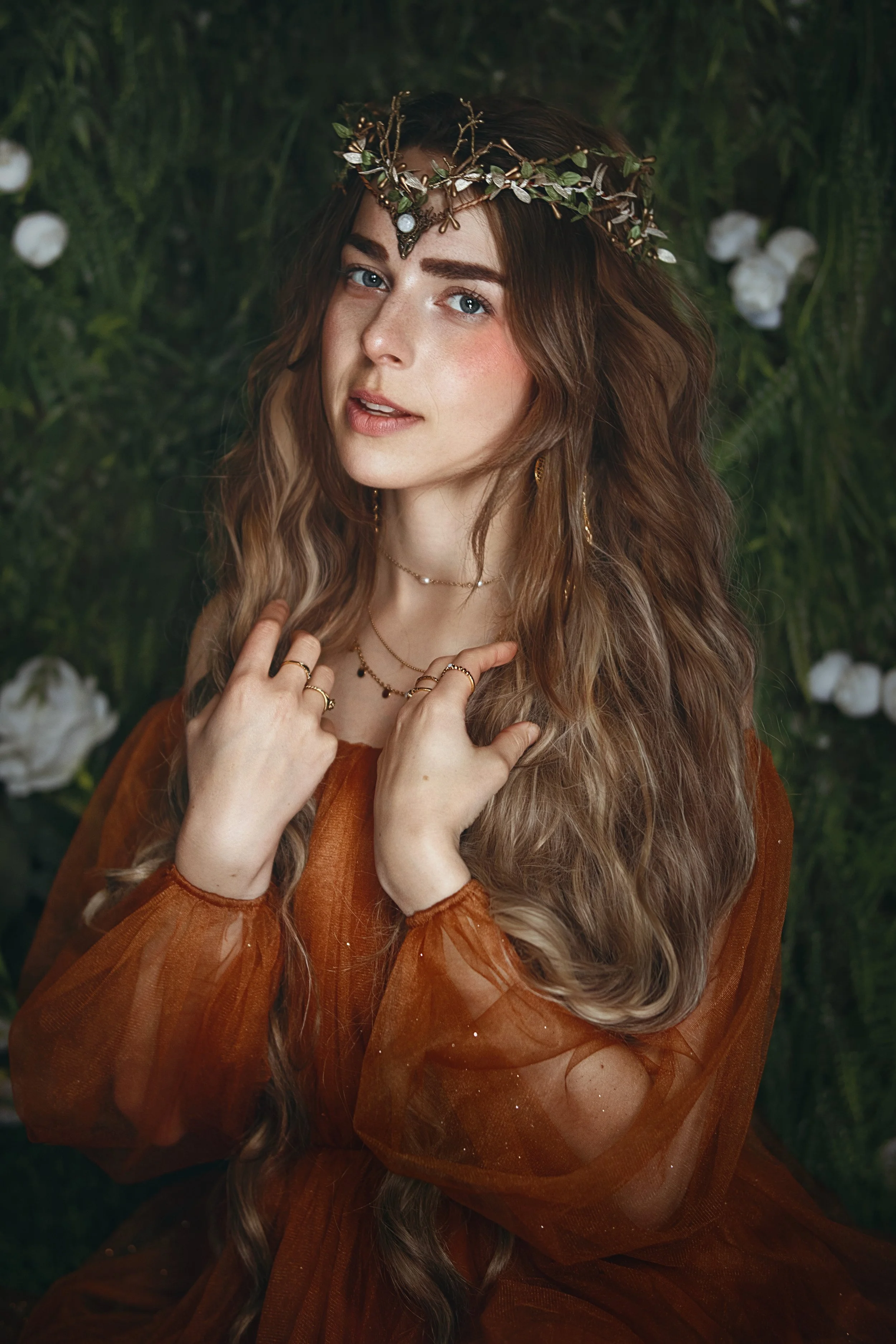 Person with long wavy hair wearing a brown dress and a floral headpiece against a dark green floral background.