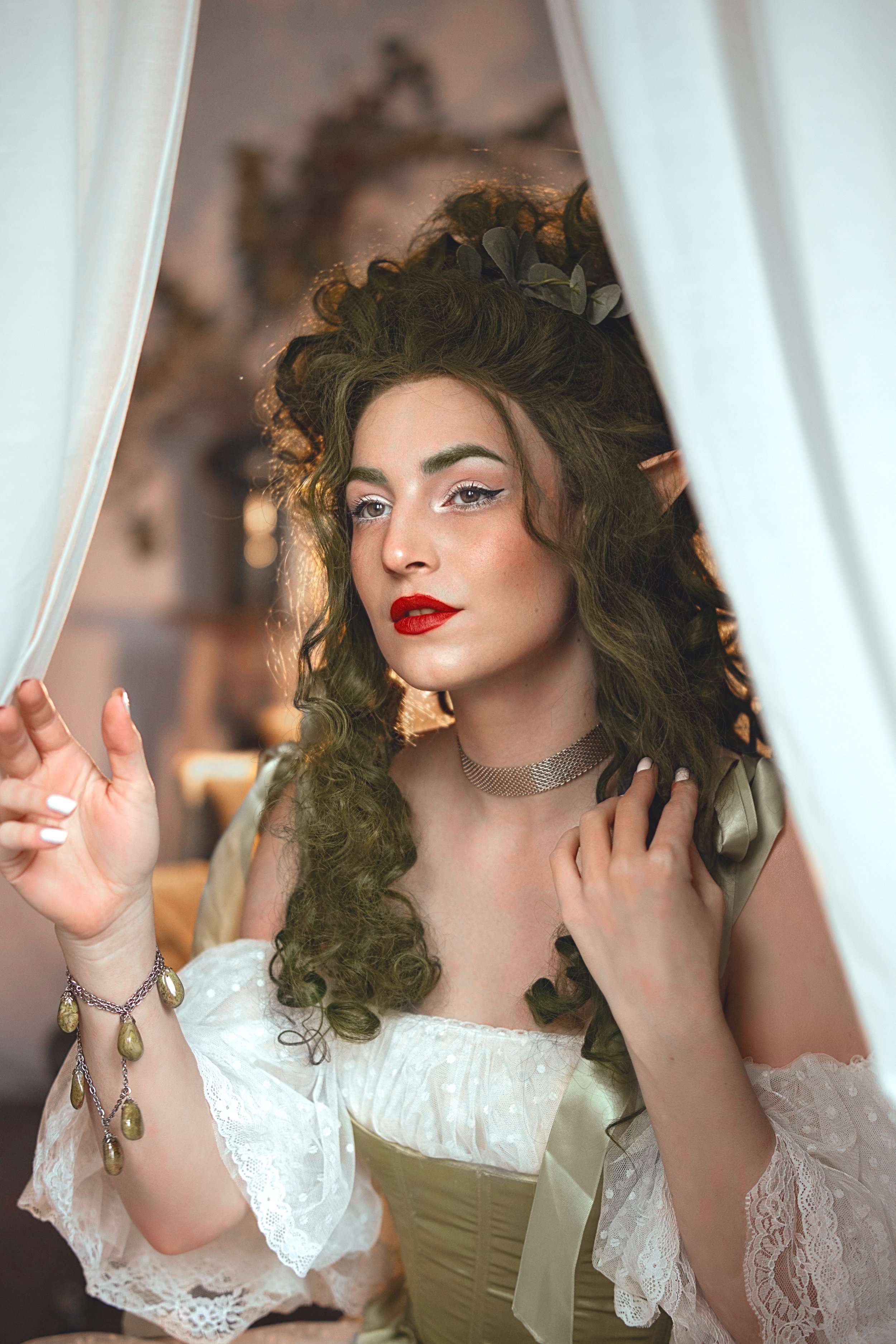Woman with elf-like features, including pointed ears and elaborate hairstyle, wearing a green and white dress with lace sleeves and accessories, peering through white curtains.