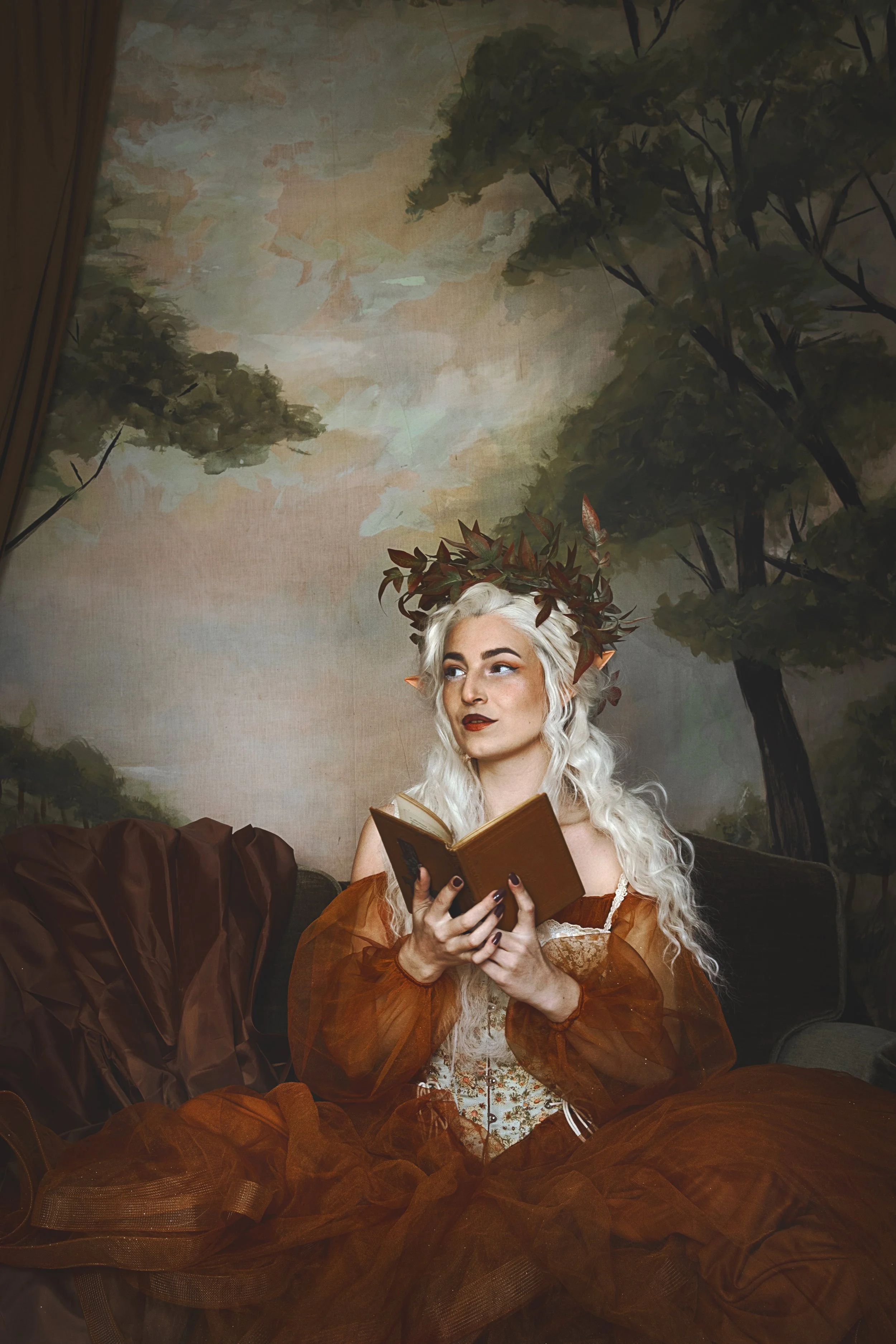 Woman dressed in a whimsical, fantasy-style costume with a crown of leaves, holding an open book, seated in front of a painted forest backdrop.