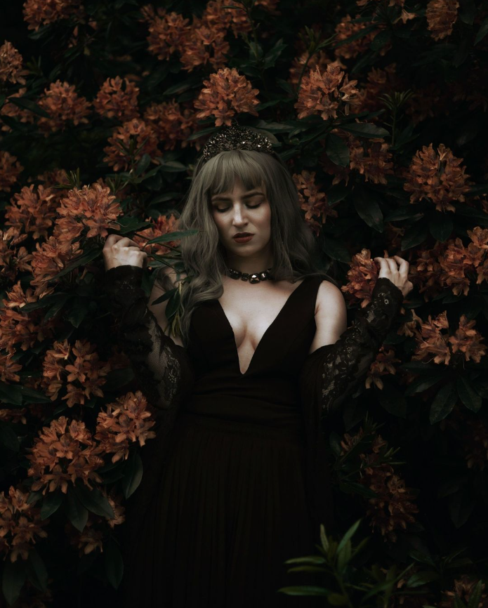 Person in a dark dress standing in front of large, lush green bushes with vibrant orange flowers.