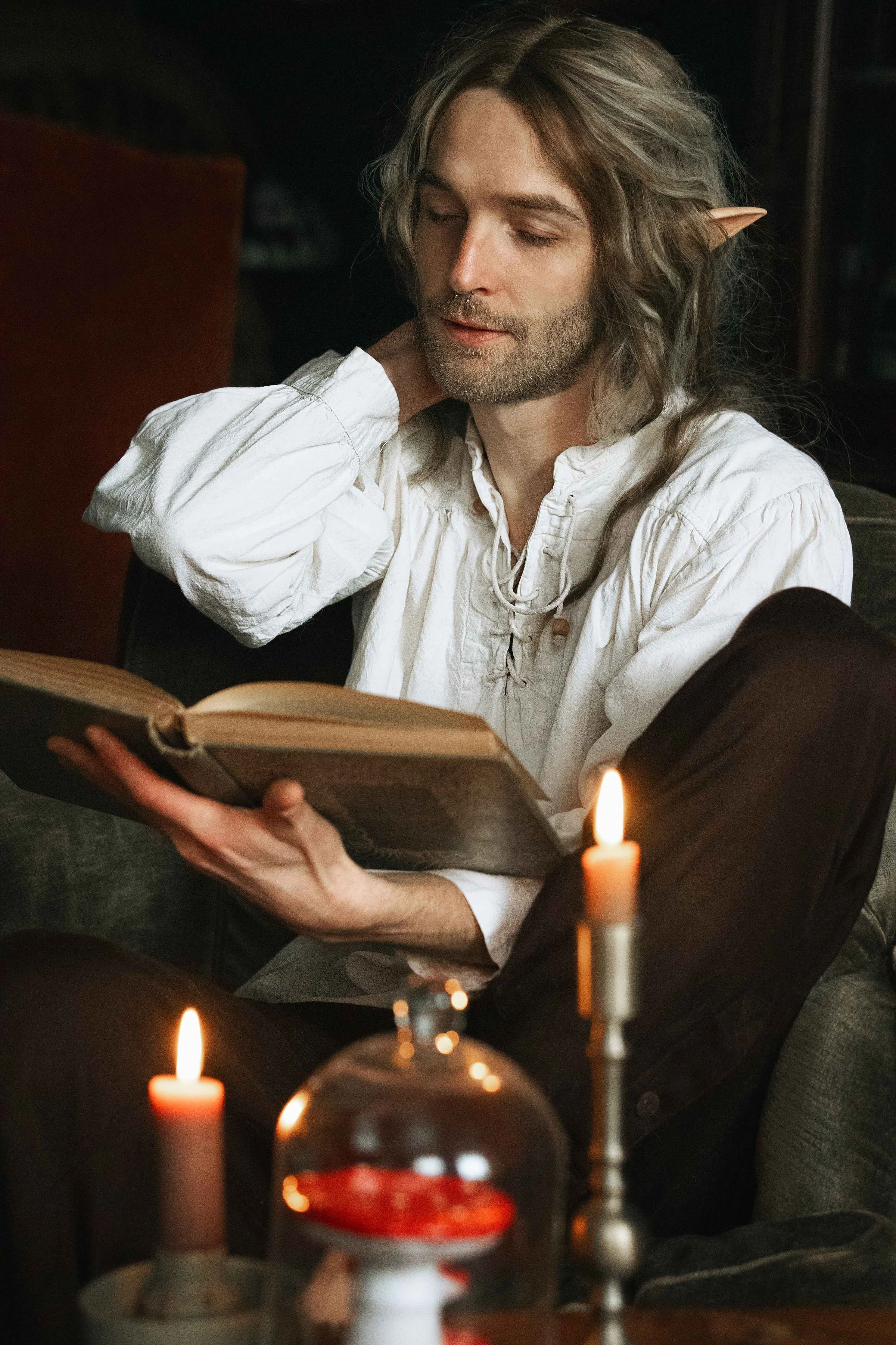 Dreamy fine art portrait of Rico as a scholar reading by candlelight. Painterly fantasy photography inspired by folklore and classical paintings, featuring books, glass bottles and warm light. Moody, romantic storytelling imagery in a fine art studio