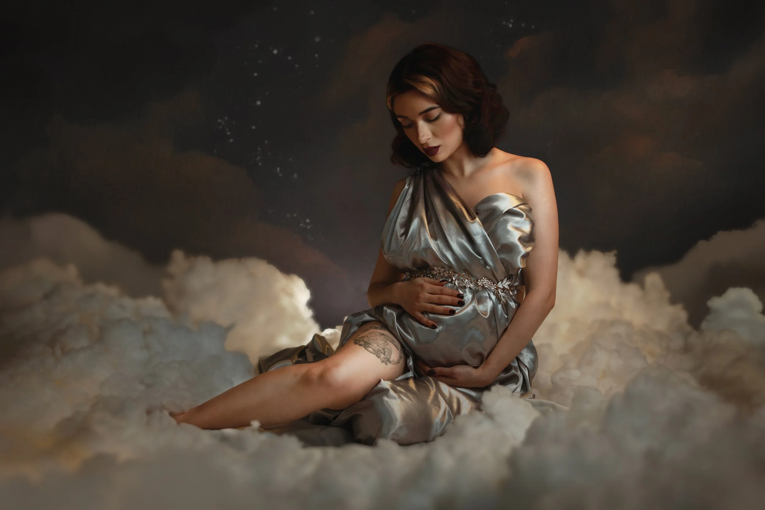 Dreamy fine art maternity portrait of a pregnant woman surrounded by clouds and soft golden light. Ethereal pregnancy photography inspired by classical paintings, divine femininity and mythology. Romantic, painterly motherhood imagery with flowing fa