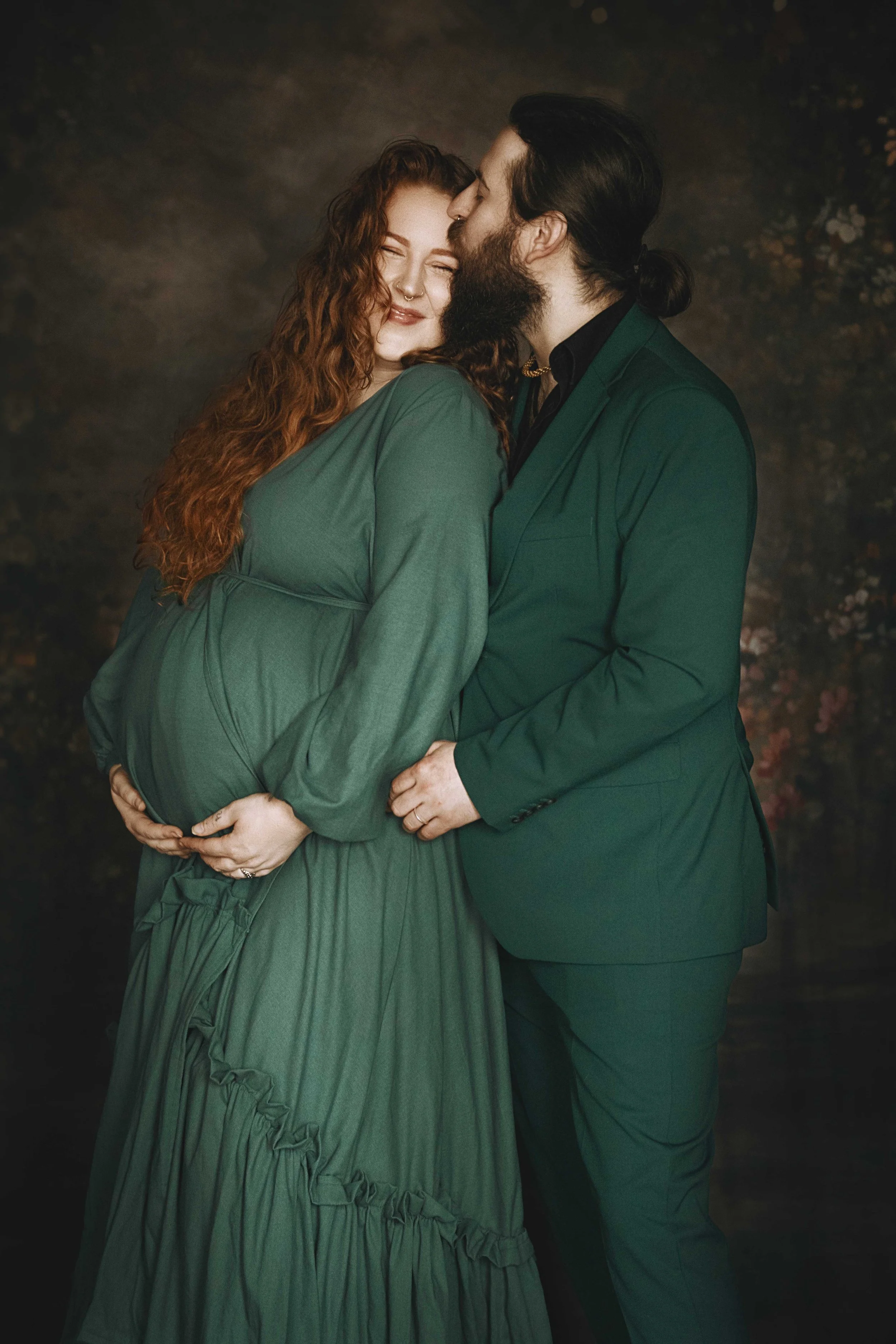 Pregnant woman in green dress embraced by man in green suit, posing affectionately with dark background.