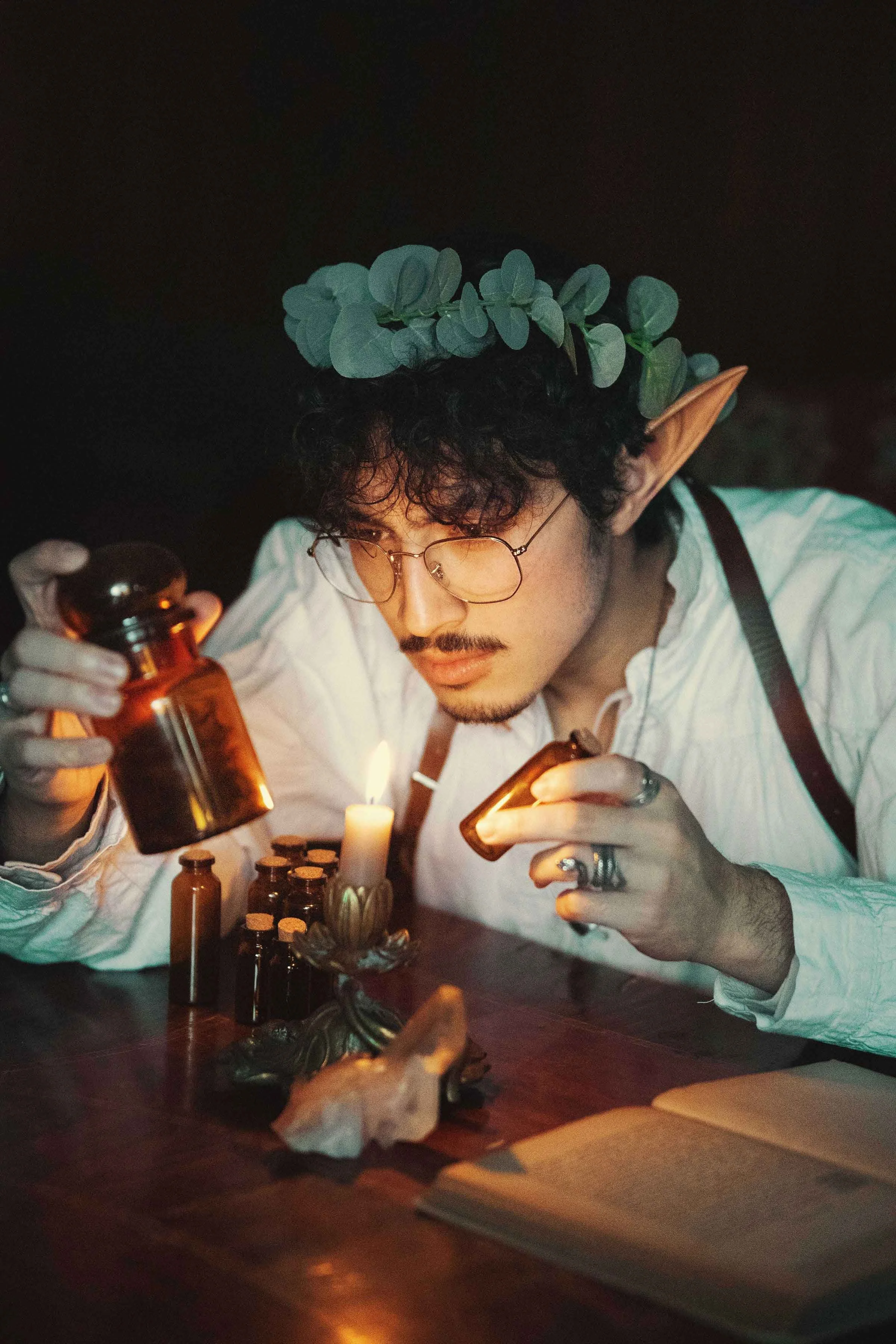 Dreamy fine art portrait of Dewa as a folkloric alchemist, surrounded by glass bottles, herbs and candlelight. Painterly fantasy photography inspired by historical apothecaries and storytelling portraiture. Studio shoot in the Netherlands.