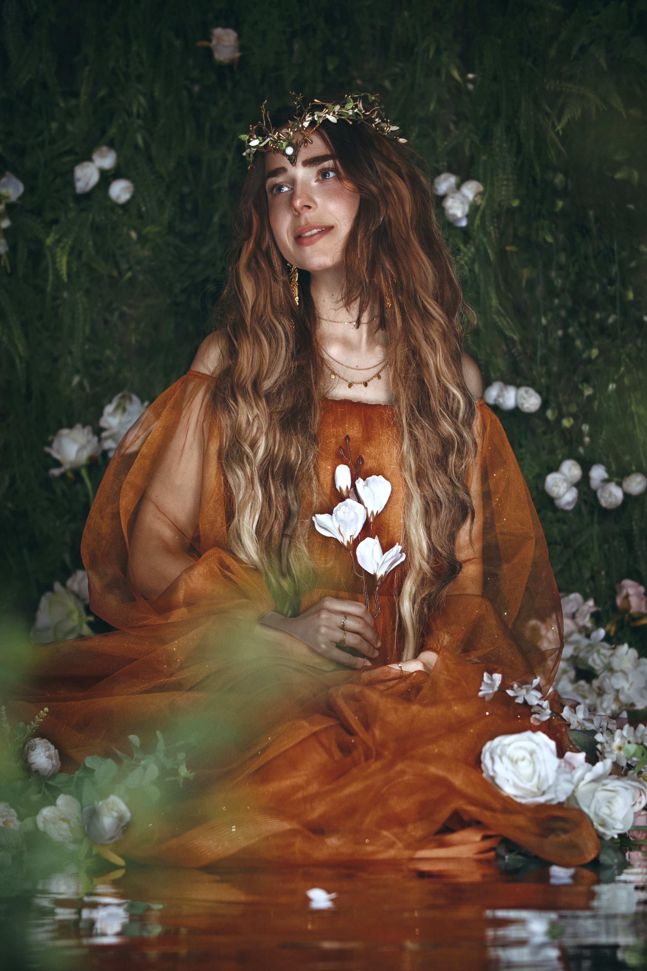 Woman in orange dress with flower crown, holding white flowers, surrounded by greenery and white roses.