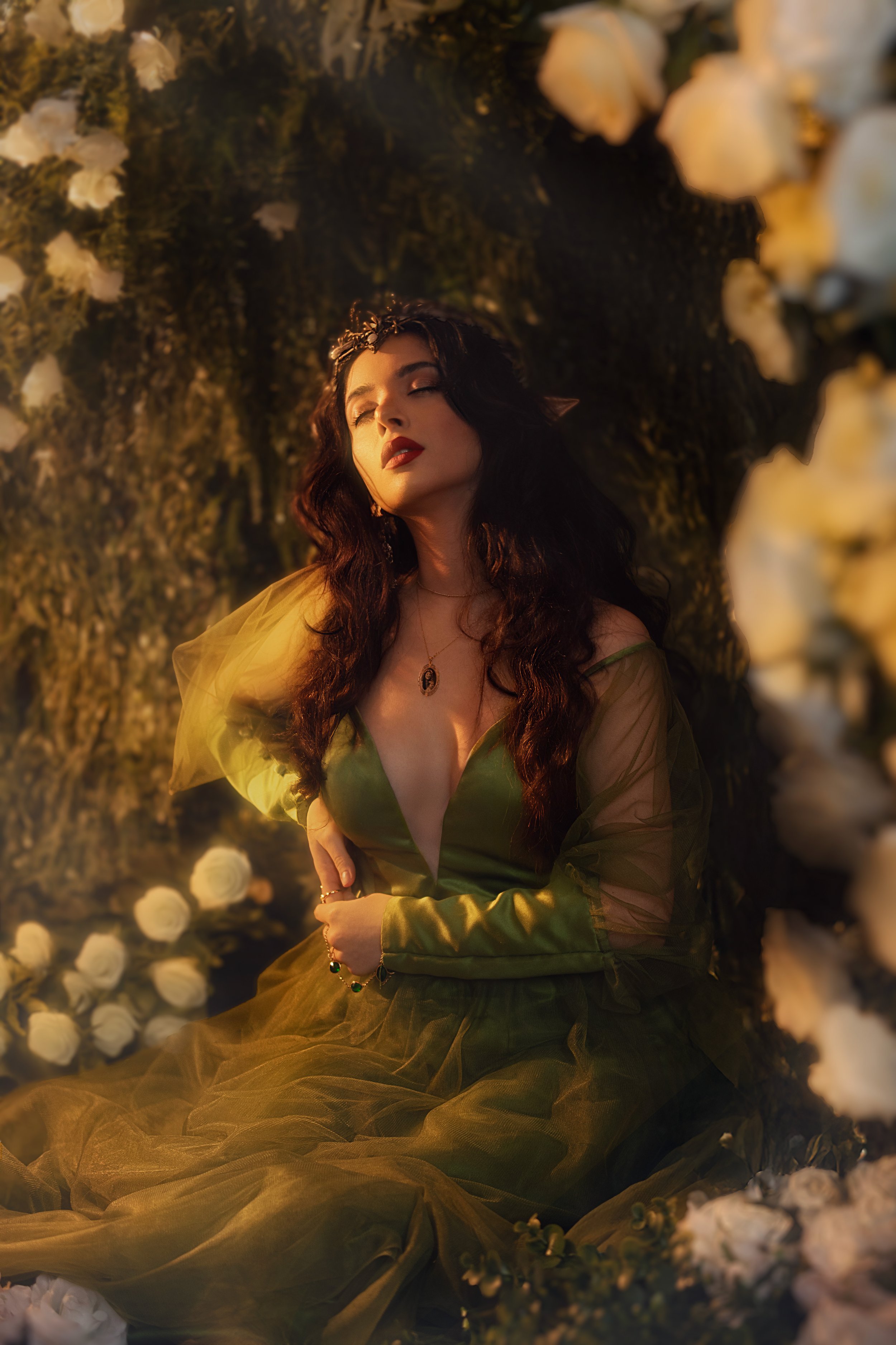 Woman in a green dress with long dark hair, wearing a golden crown, surrounded by white roses and greenery, with eyes closed in a serene pose.