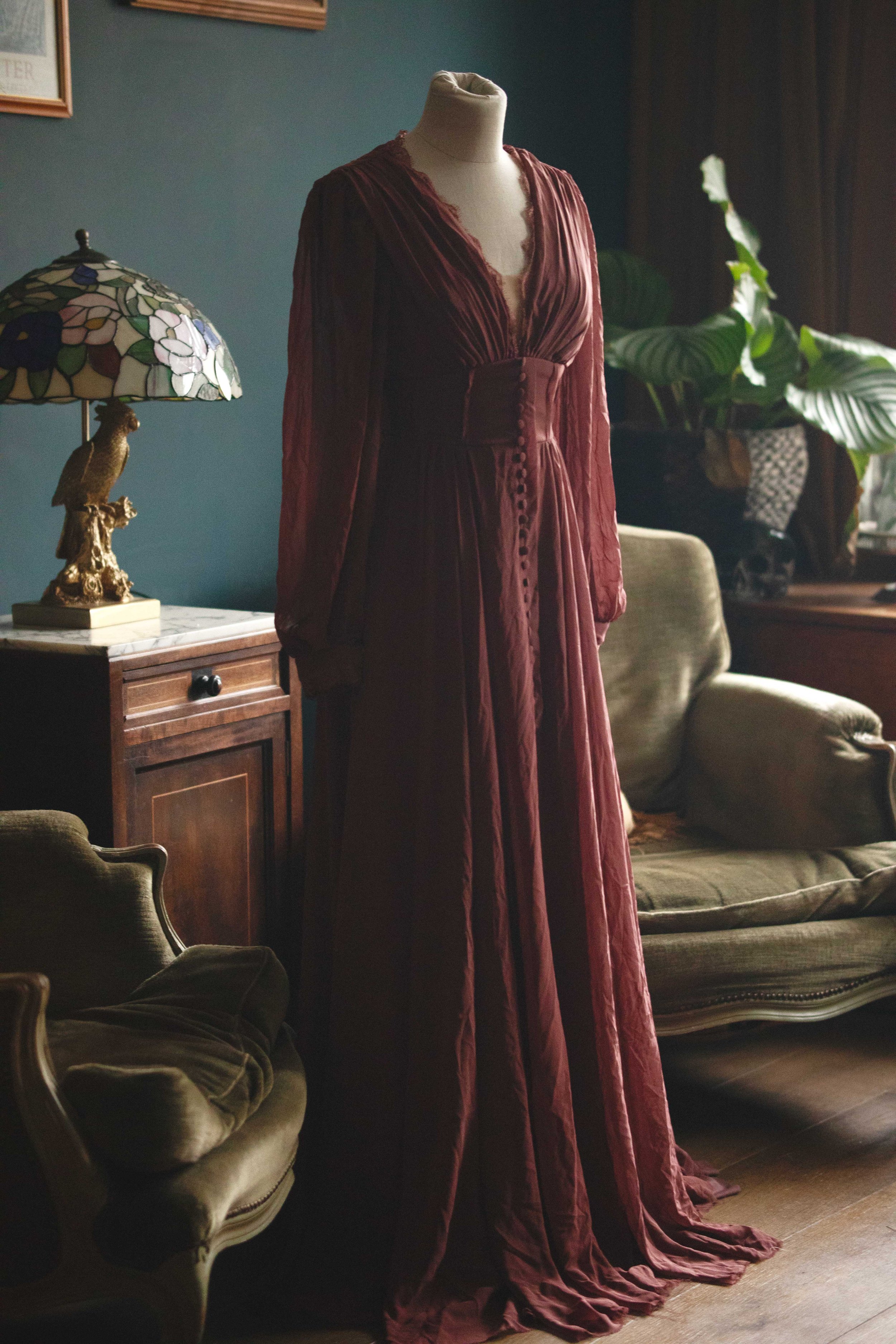 Romantische mauve jurk met lange mouwen, diepe V-hals en een brede tailleband met beklede knoopjes over de hele lengte. De zachte, vloeiende stof geeft een elegante, bijna middeleeuwse uitstraling. XS - S