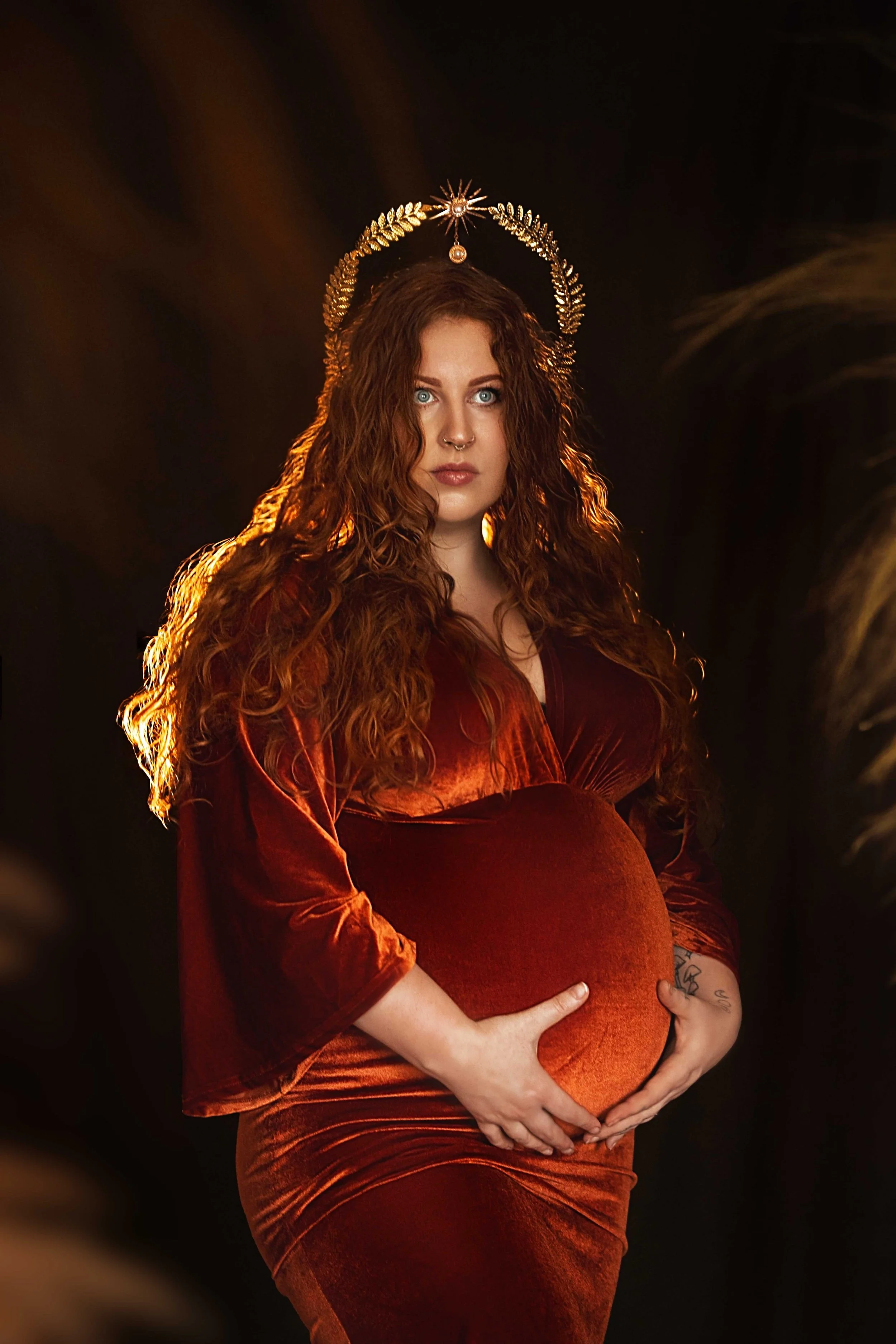 Pregnant woman with long red hair wearing a crown and rust-colored dress, holding her belly.