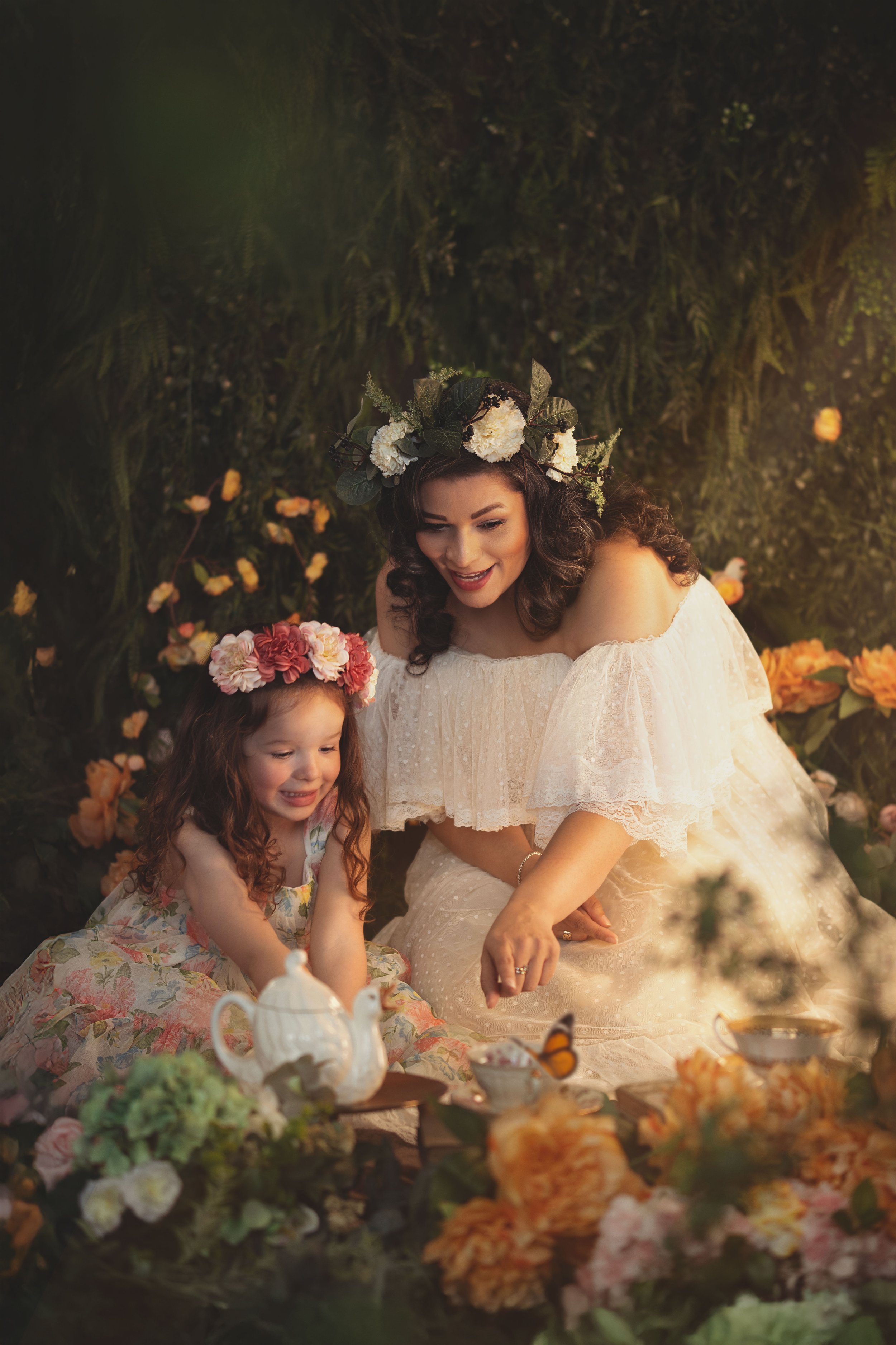 Mother and daughter in floral crowns having a tea party in a garden setting with flowers and a teapot.