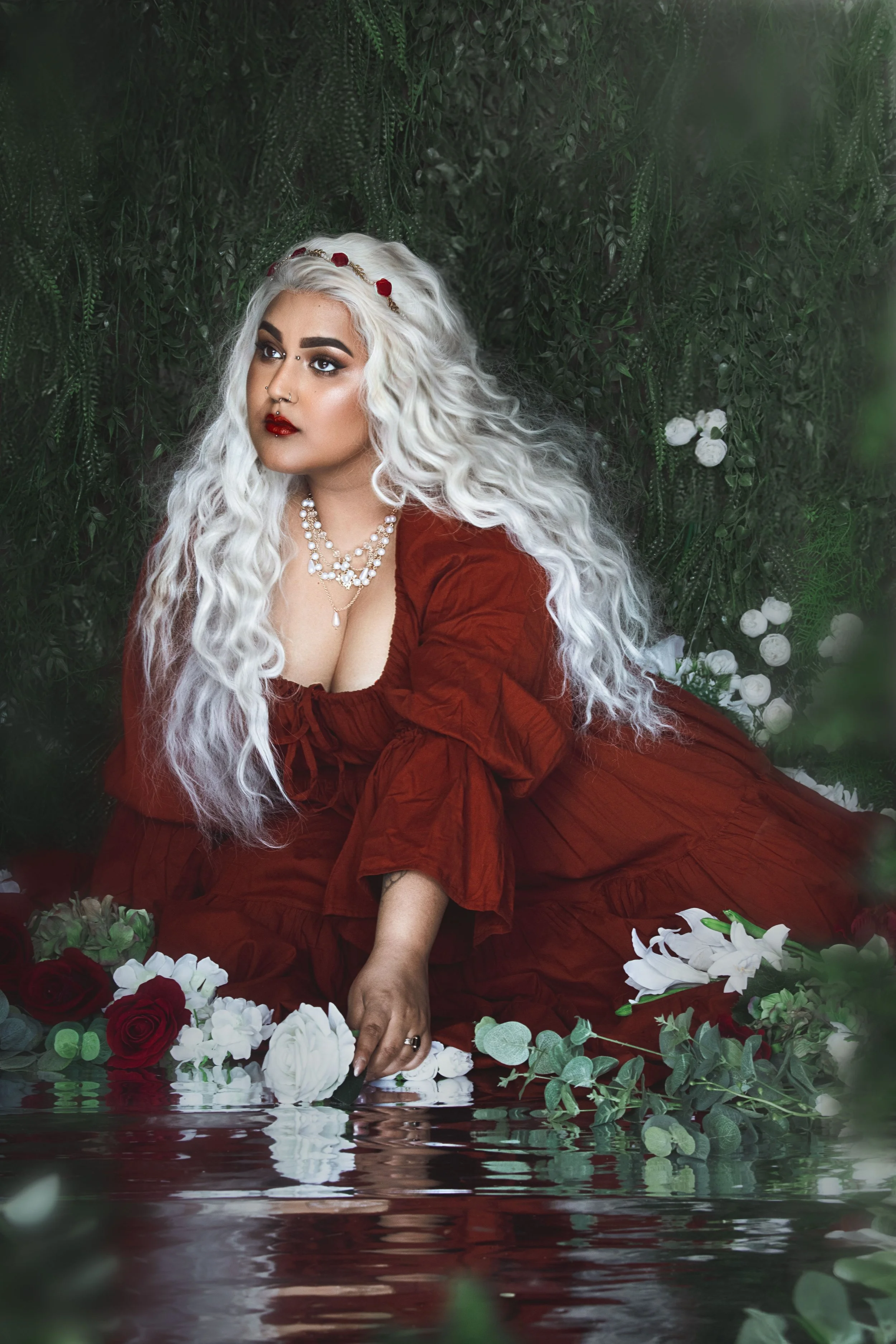 A person with long white hair dressed in a red gown, sitting by a reflective water surface surrounded by flowers and greenery, gently touching the water.